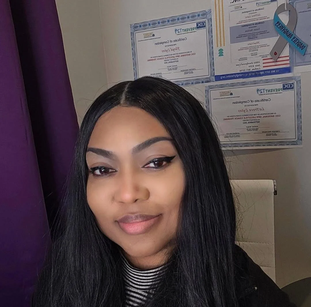 A woman with long black hair and makeup, posing indoors near purple curtains, with framed certificates or documents on the wall behind her.