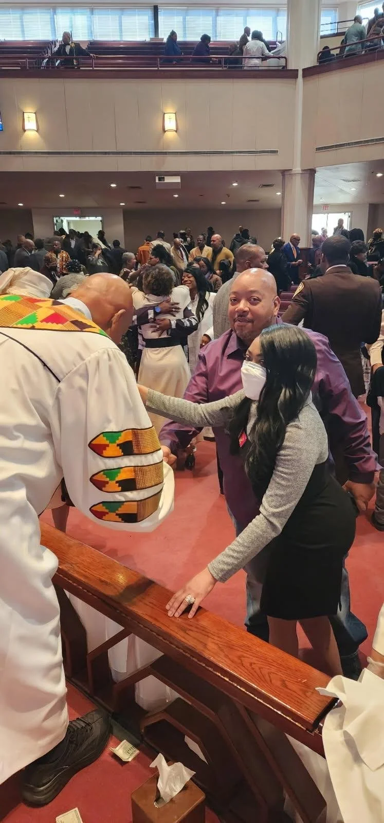 A diverse group of people gathered in a church or community hall, engaging in conversation and greetings, with some individuals shaking hands and embracing.