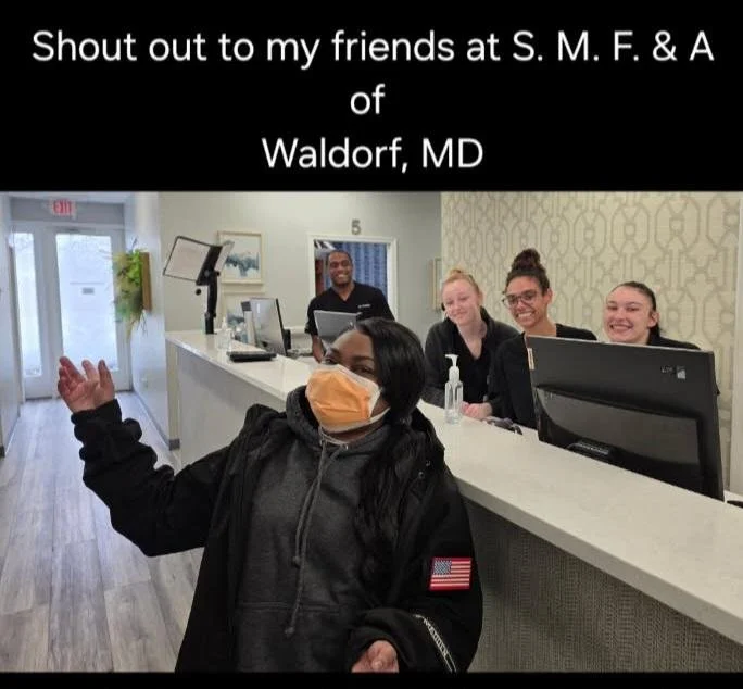 Four smiling reception staff behind a counter with a woman in front waving, in a reception area of a medical or dental office, with a sign that reads 'Shout out to my friends at S. M. F. & A of Waldorf, MD'.