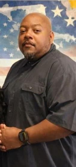 A man with a shaved head and beard wearing a black shirt, standing in front of an American flag-themed background.