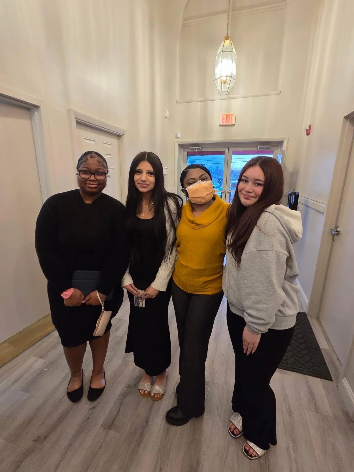 Four women standing together indoors, smiling at the camera. One woman is wearing a black dress and glasses, another in a white sweater and black skirt, the third in a yellow sweater and black pants wearing a face mask, and the fourth in a grey hoodi