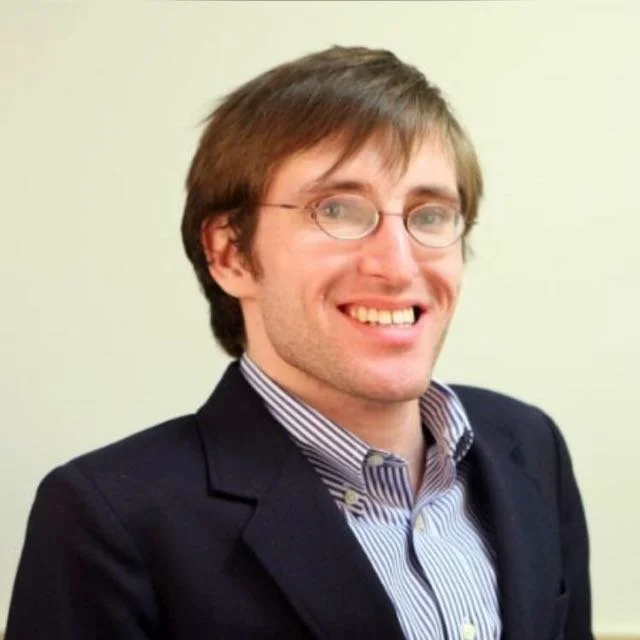 A young man with glasses, light brown hair, wearing a dark suit jacket over a striped shirt, smiling at the camera.
