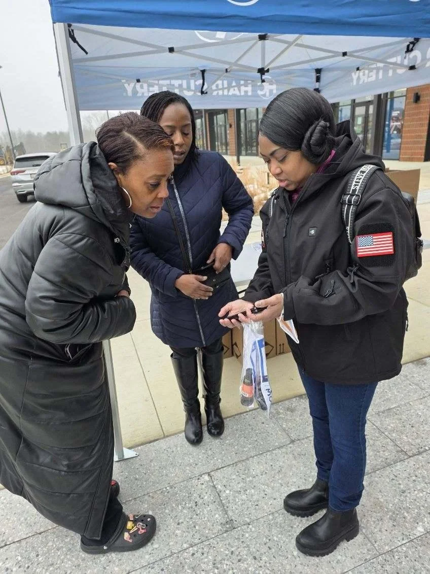Three women standing outside under a blue tent, looking at a phone. One woman is showing something on her phone to the other two. They are wearing jackets, and one has an American flag patch on her sleeve. There are cars and a building in the backgro