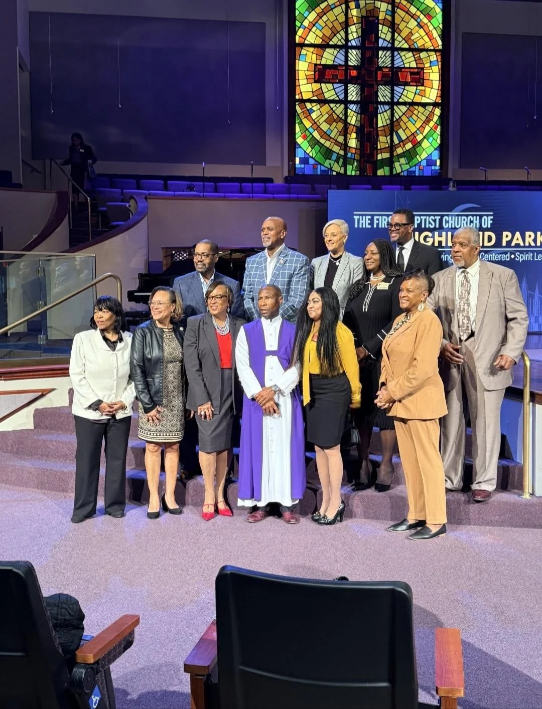 A group of church leaders and members posing together in sanctuary with stained glass cross window in the background, at The First Baptist Church of Highland Park.