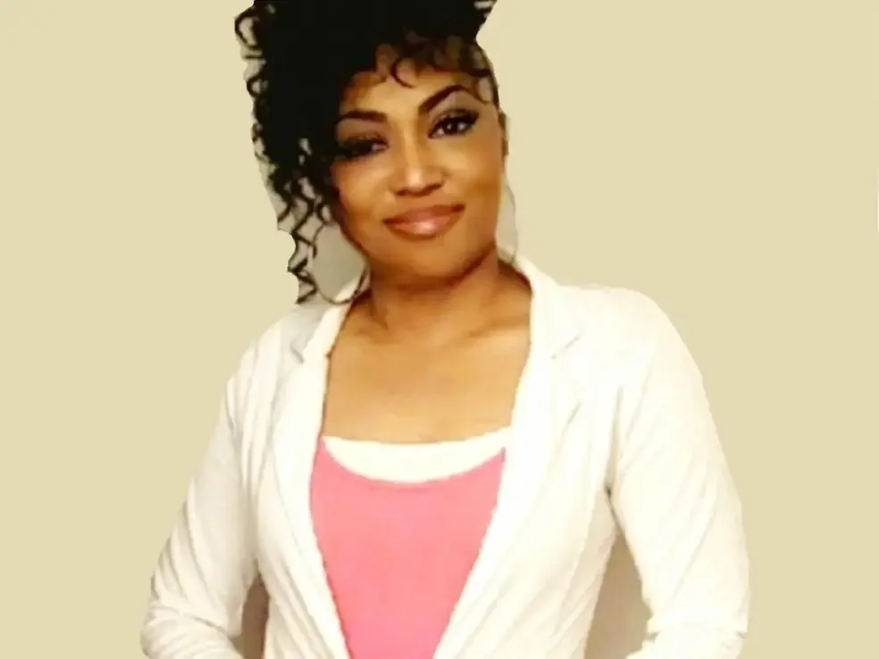 A woman with dark curly hair, wearing a white jacket over a pink top, smiling at the camera.
