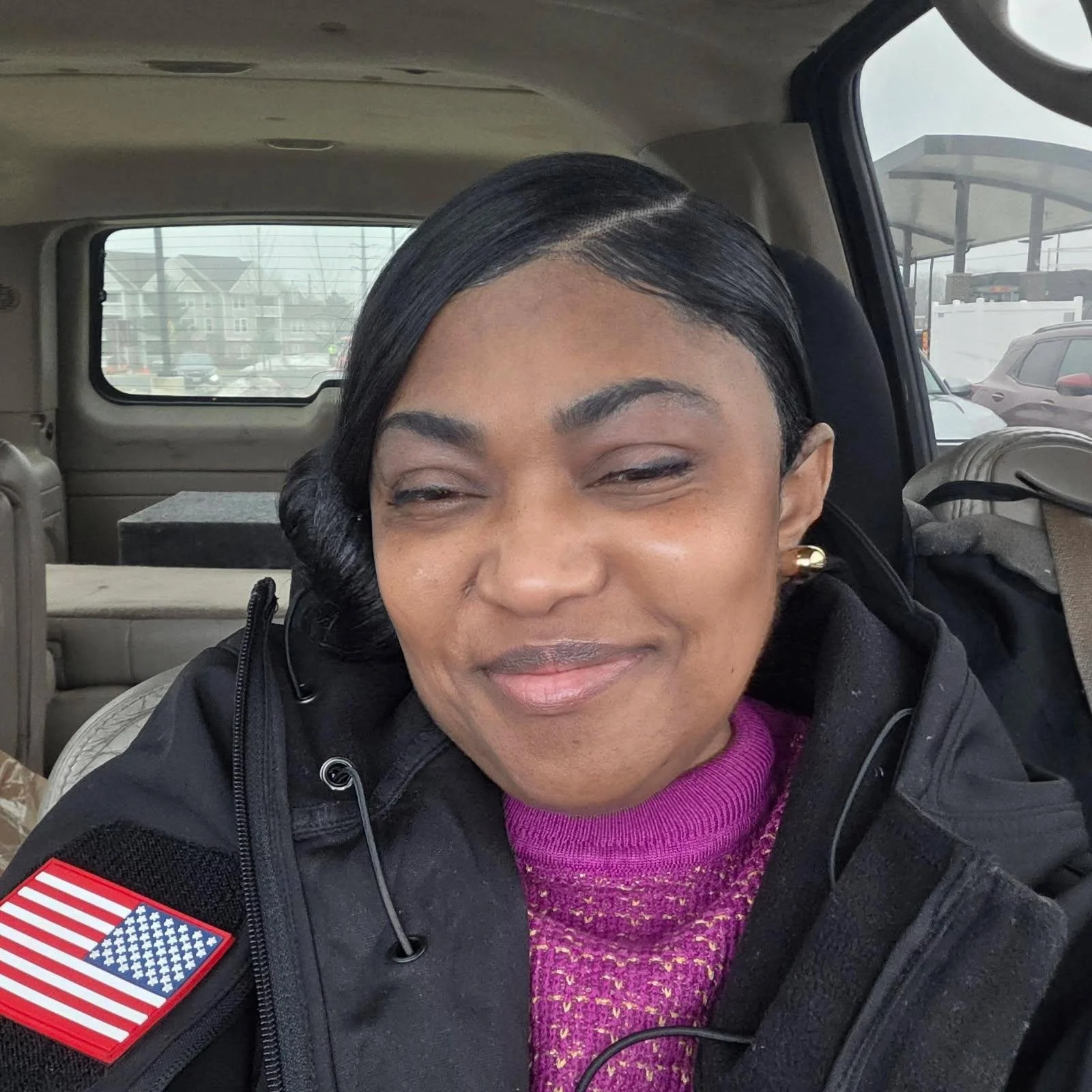 A woman with dark hair styled in waves, smiling inside a vehicle, wearing a purple sweater, black jacket with an American flag patch on the sleeve, gold earrings, and earbud cords visible.