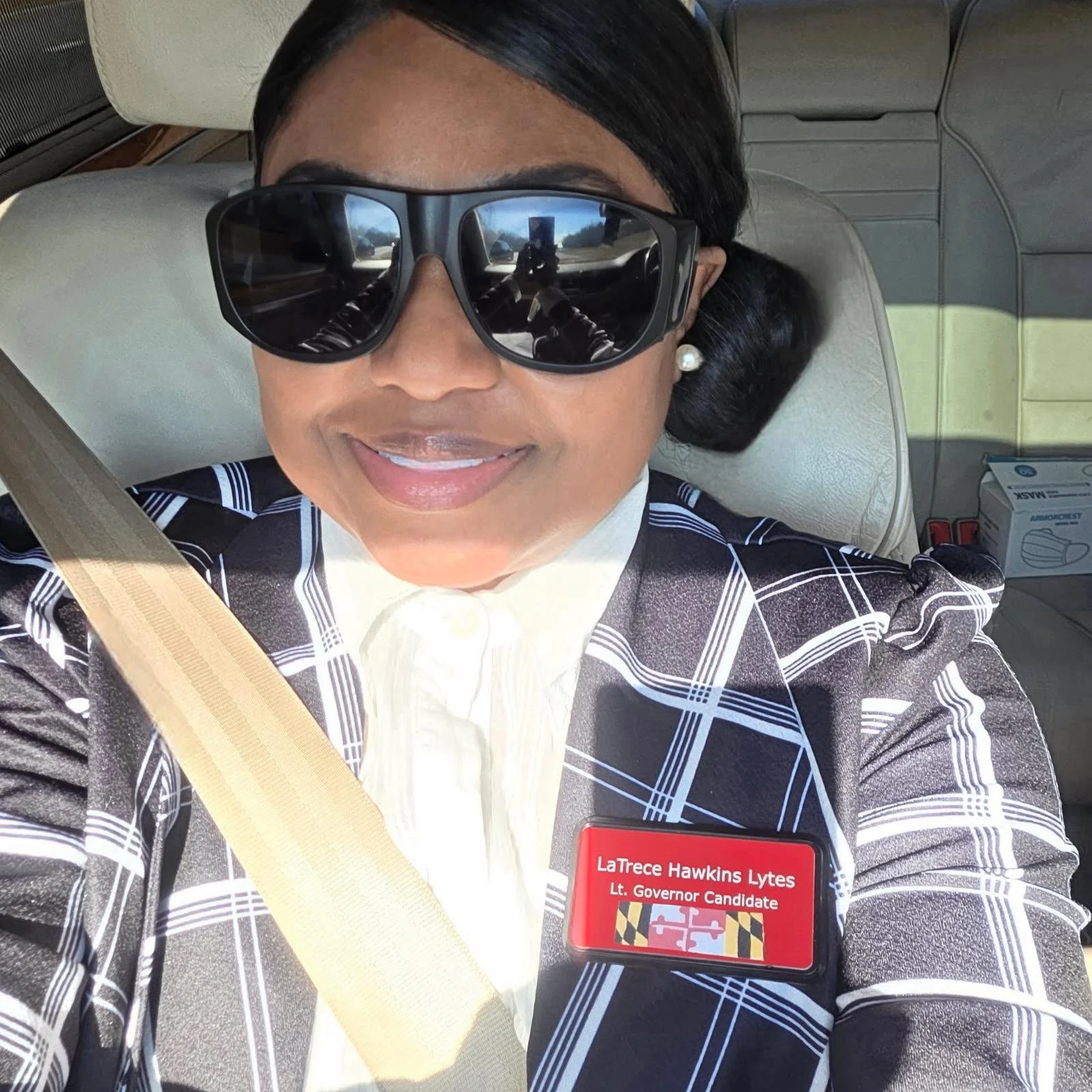 Woman wearing large sunglasses and pearl earrings, smiling while sitting in a car. She is wearing a plaid blazer with a name badge that reads "LaTrece Hawkins Lytes, Lt. Governor Candidate."