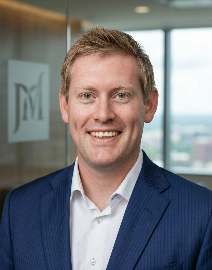 A smiling man in a navy blue suit and white shirt standing in a modern office with large windows and a city view.