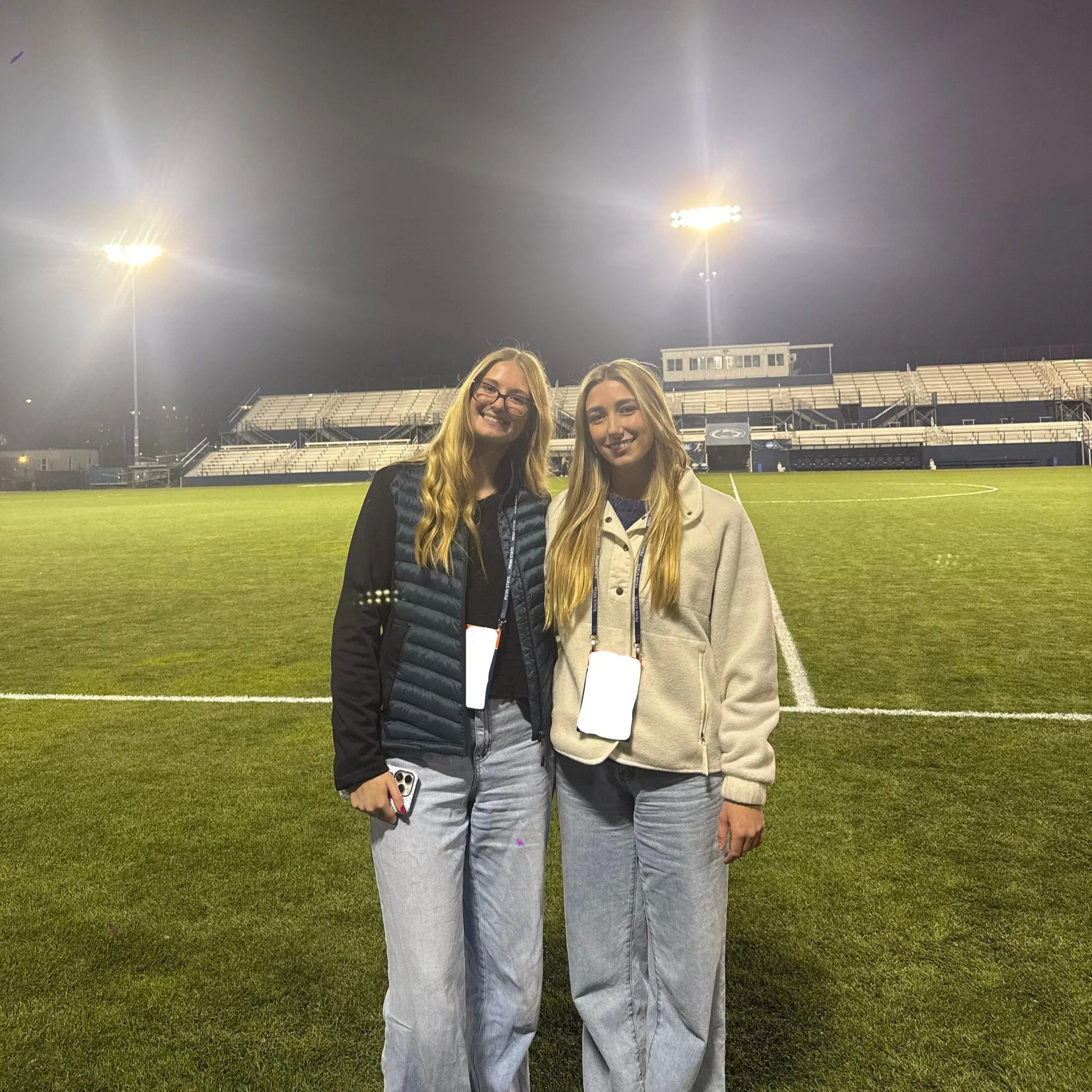 Two young women standing on a football field at night, smiling, with empty bleachers and stadium lights in the background.