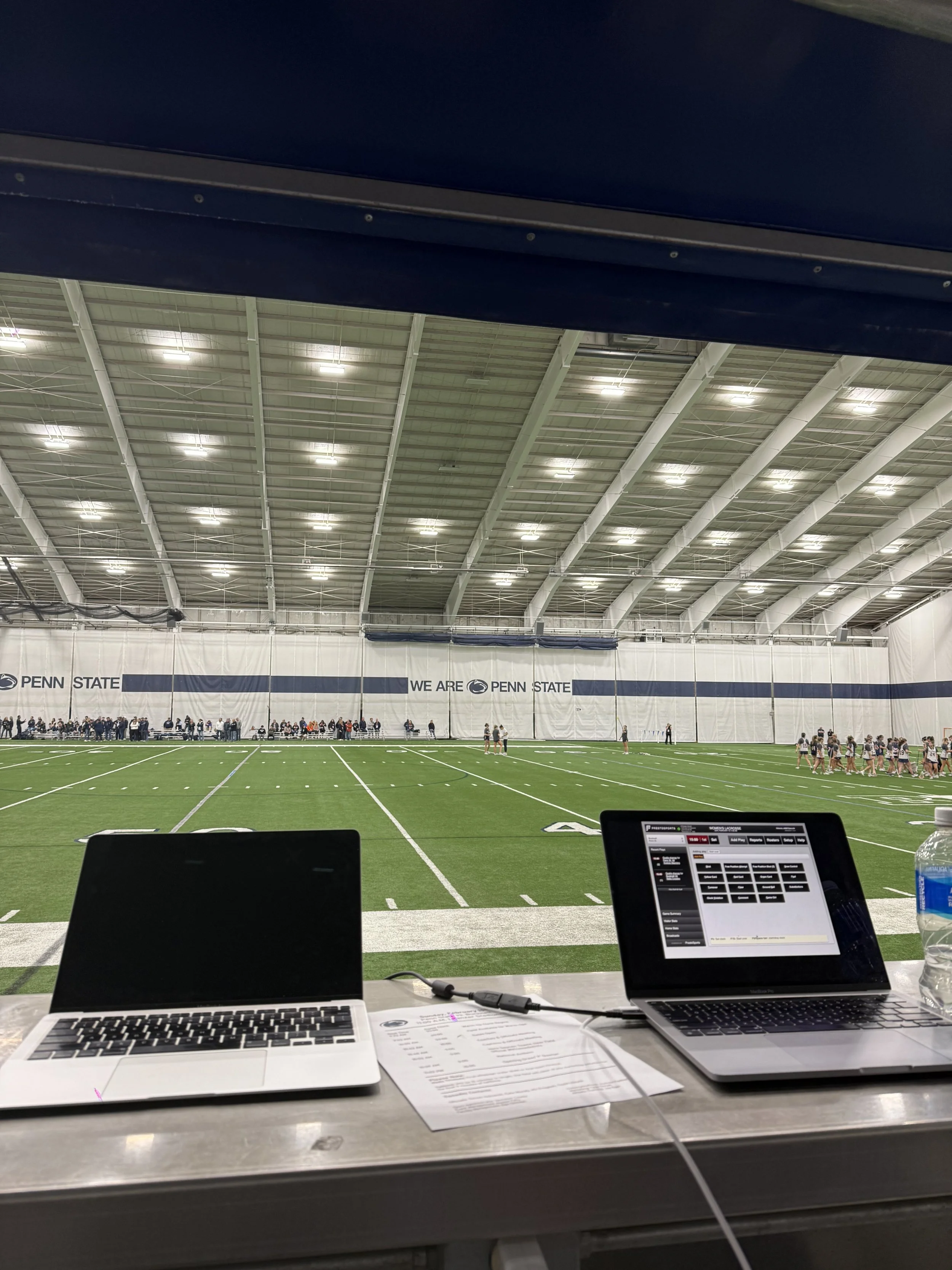 Inside a large indoor football facility at Penn State, with players practicing on the field, and two laptops and a water bottle on a table in the foreground.