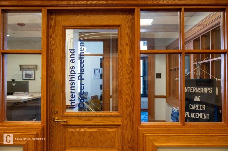 View through a wooden-framed glass door into an office with signs for internships and career placement.