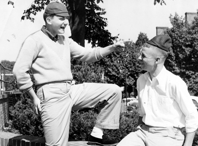 Two boys outdoors, one standing with a raised knee and pointing, the other sitting on a brick ledge, smiling at him, both wearing caps and casual clothes.