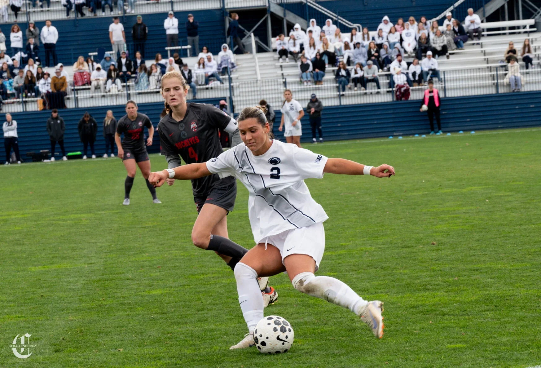 Women soccer players competing for the ball on the field during a match, with spectators seated in the background.