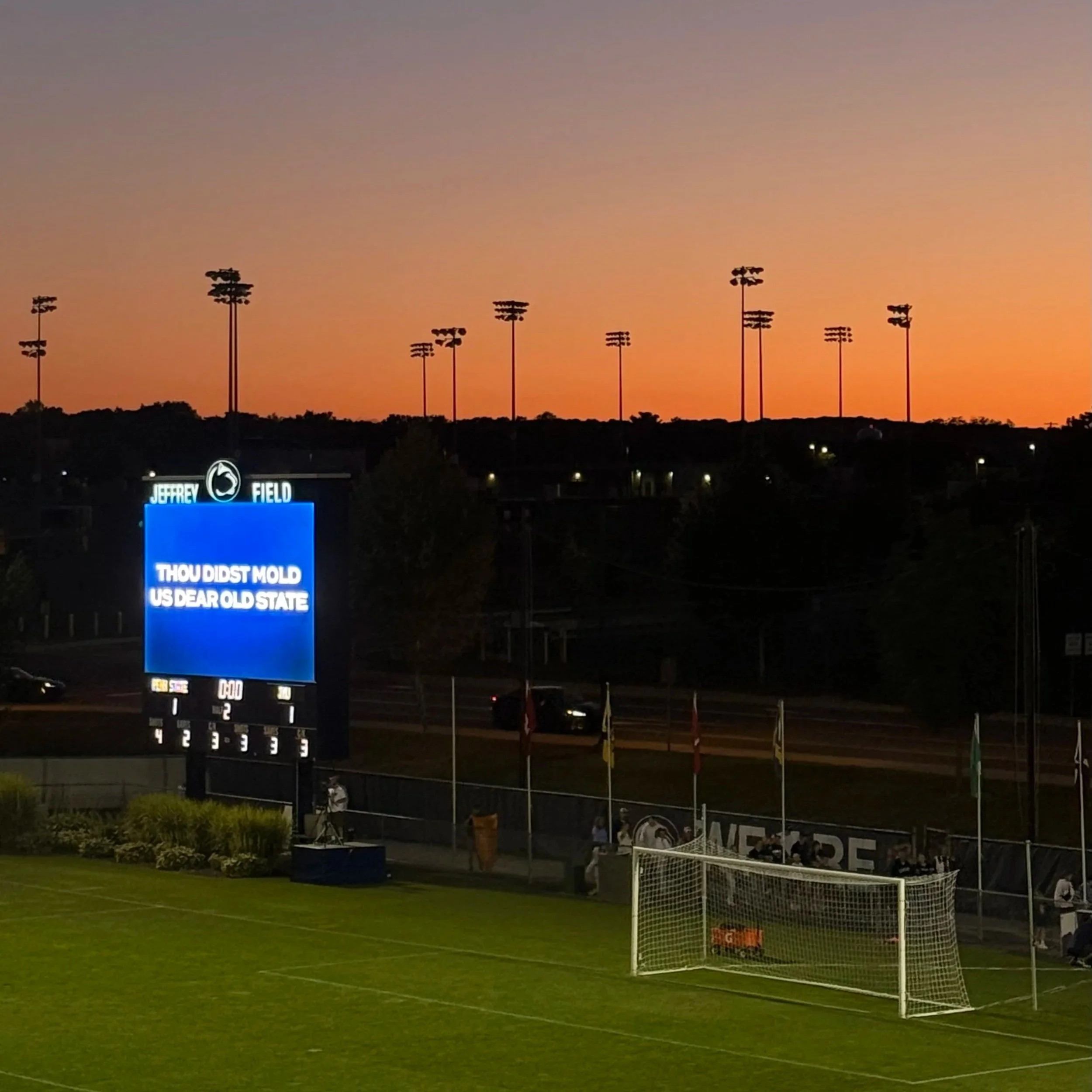 Sunset sky over a sports stadium with lights, scoreboard displaying a message, and soccer goal on a grass field in the foreground.
