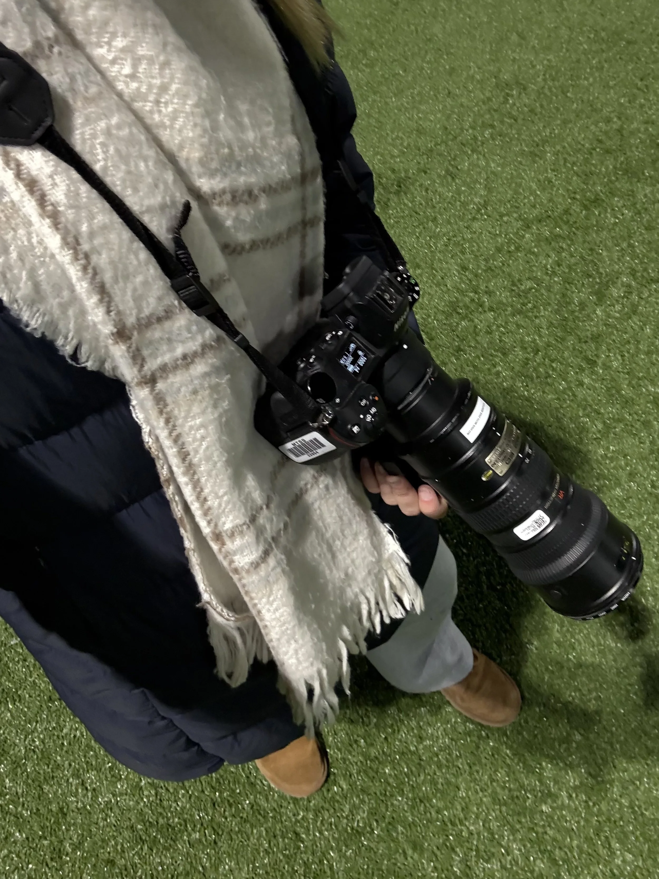Person holding a professional camera with a large telephoto lens, standing on artificial grass.