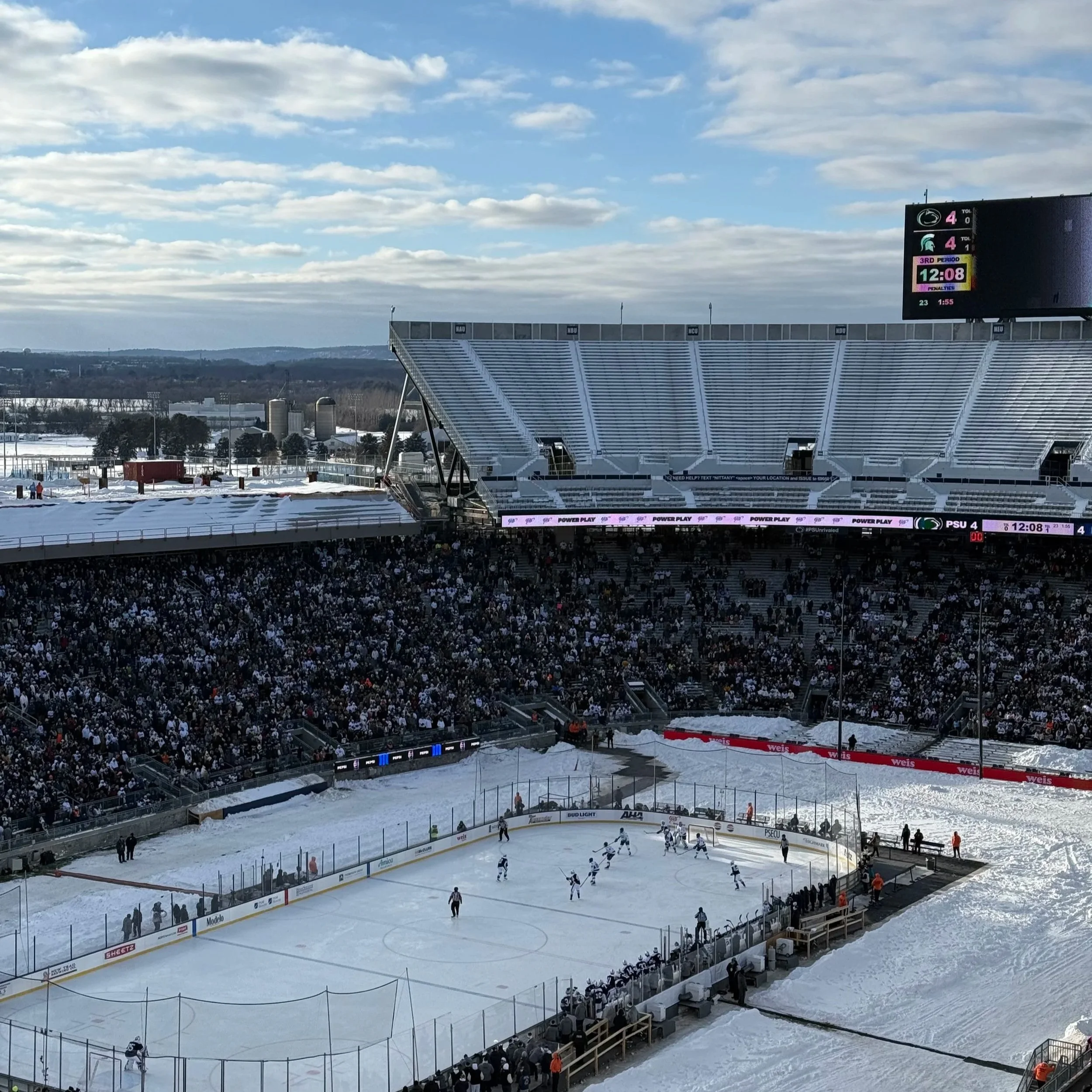 An outdoor ice hockey game being played in a snow-covered stadium with a partially filled audience watching.