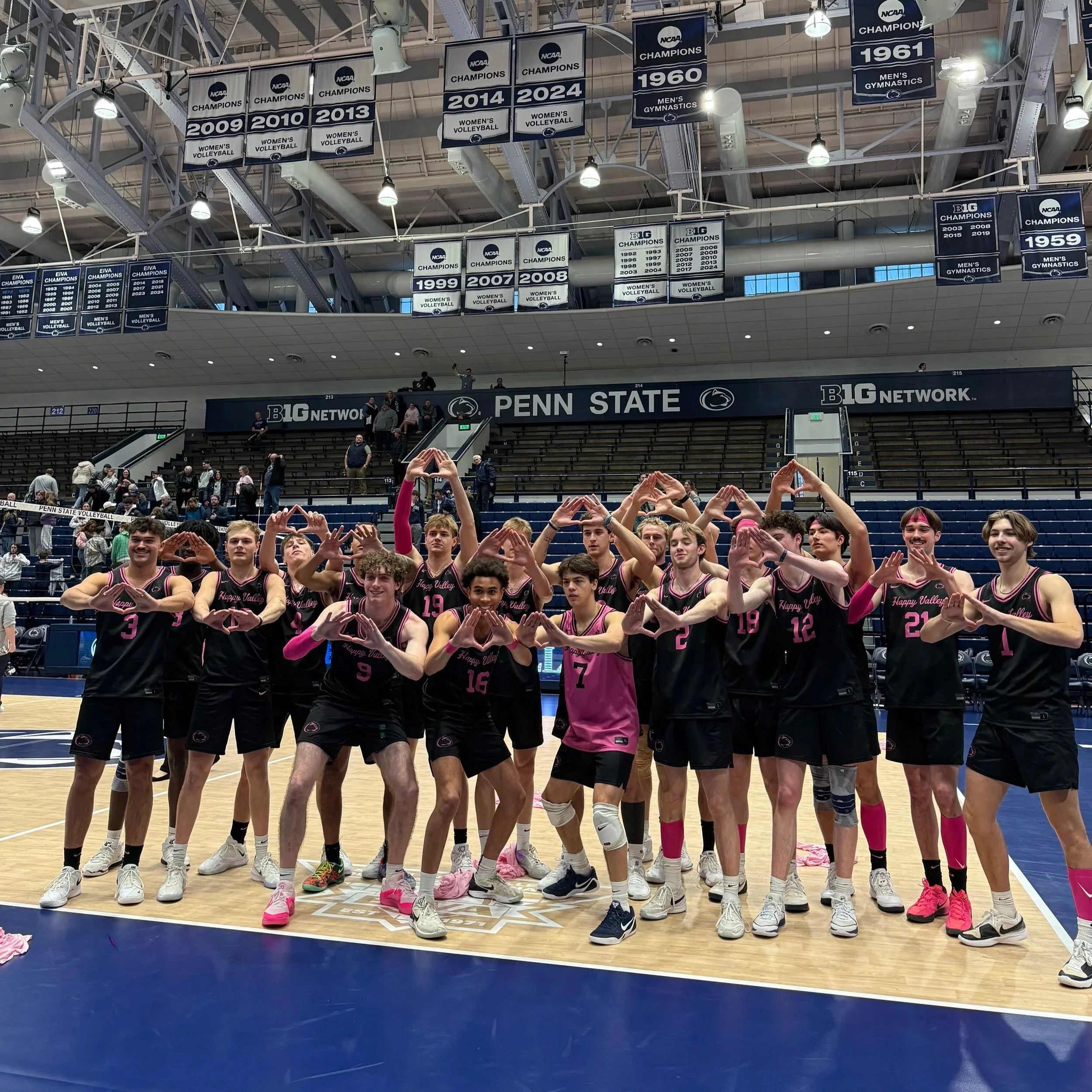 A group of young male volleyball players on an indoor court at Penn State, wearing black and pink uniforms, making heart shapes with their hands, celebrating after a match.