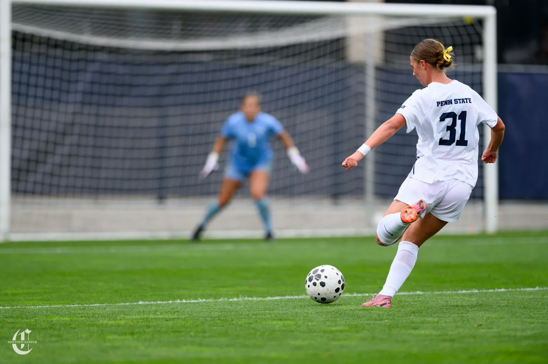 A female soccer player from Penn State taking a penalty kick, with a goalkeeper in the background preparing to save the shot, on a green soccer field.