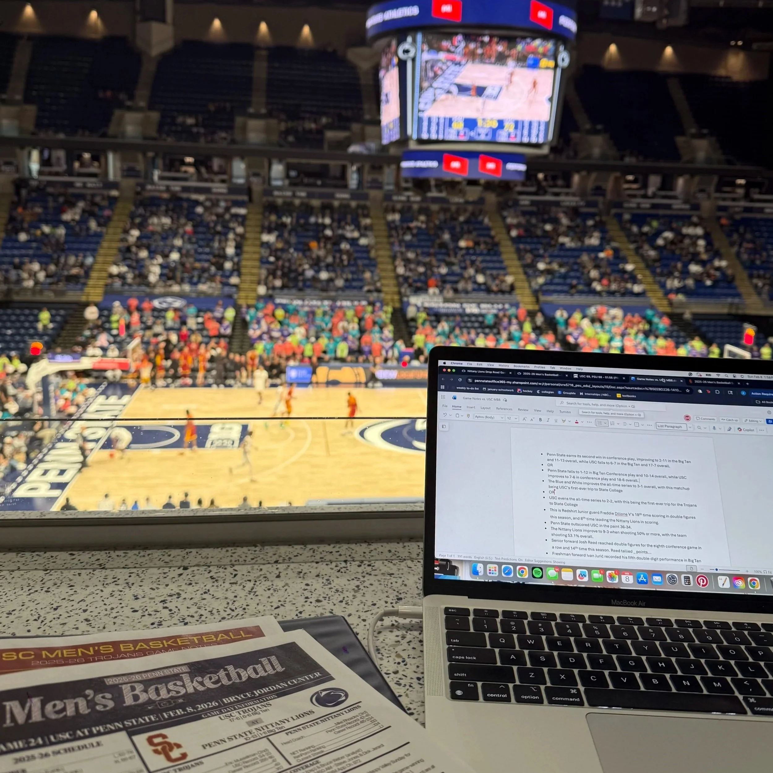 A basketball game is happening in an arena with many spectators, viewed from a press box. On the desk, there is a laptop displaying notes about the game and a printed program titled 'SC Men's Basketball.'