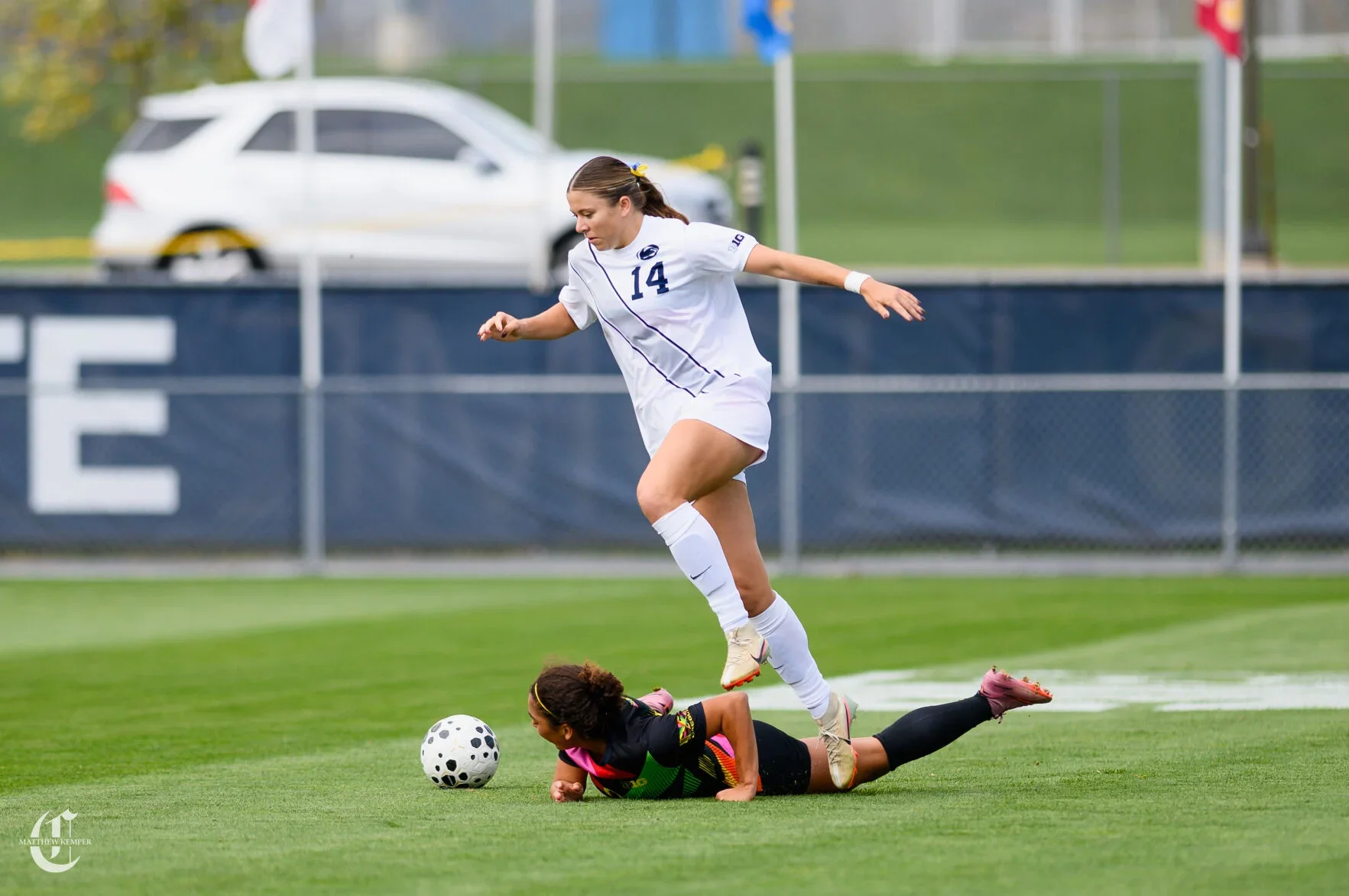 Two female soccer players competing for the ball during a match, one in a white uniform jumping over the player in black and pink on the ground