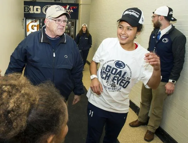 A young woman in a white Penn State soccer jersey and cap smiles and talks to an older man in a navy jacket and cap in a hallway. Other people are in the background near vending machines.