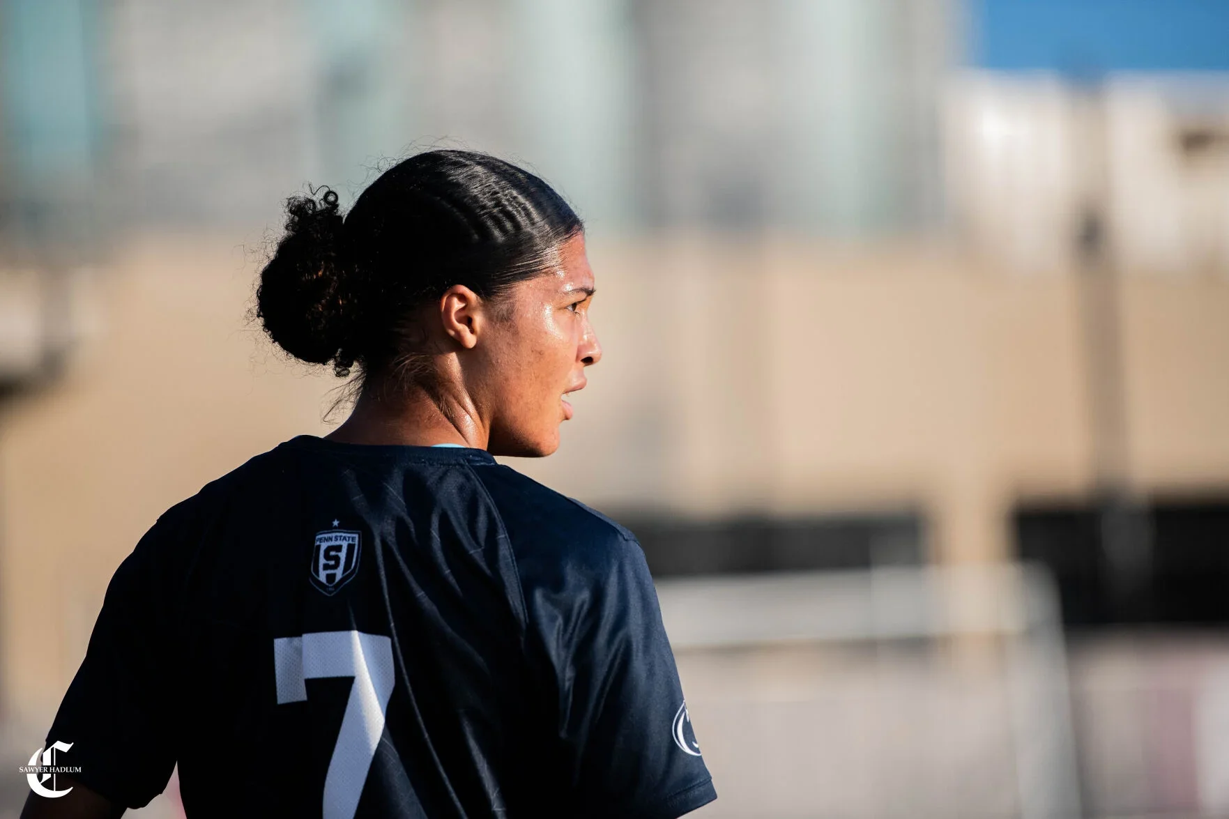 Profile of a female athlete with dark braided hair, wearing a black sports jersey with the number 7, on a sports field, with a blurred background of buildings and stadium lights.