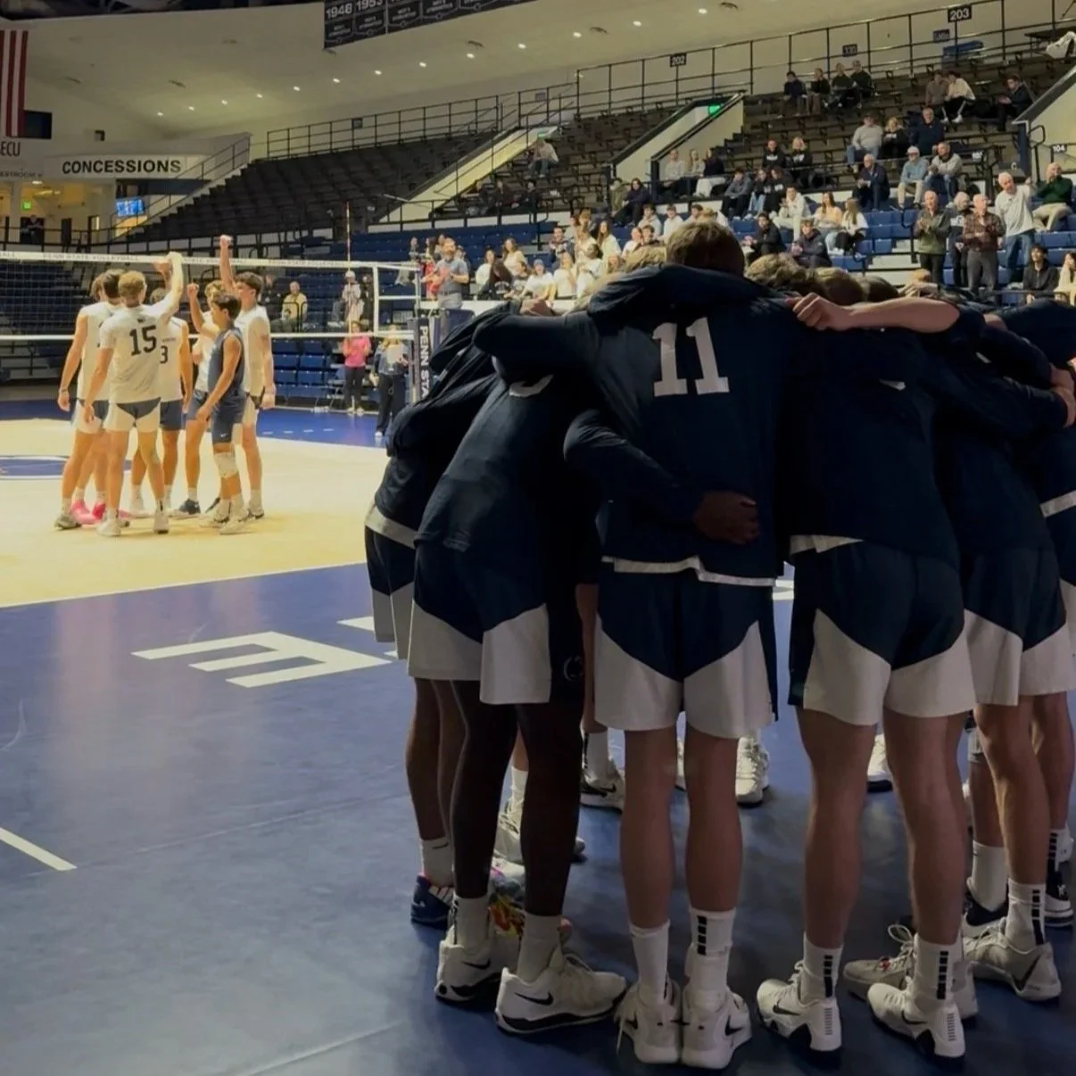 A volleyball team huddles together on the court during a game, with players wearing navy and white uniforms. In the background, their opponents, wearing white uniforms, are gathered near the net, and spectators sit in the stands.