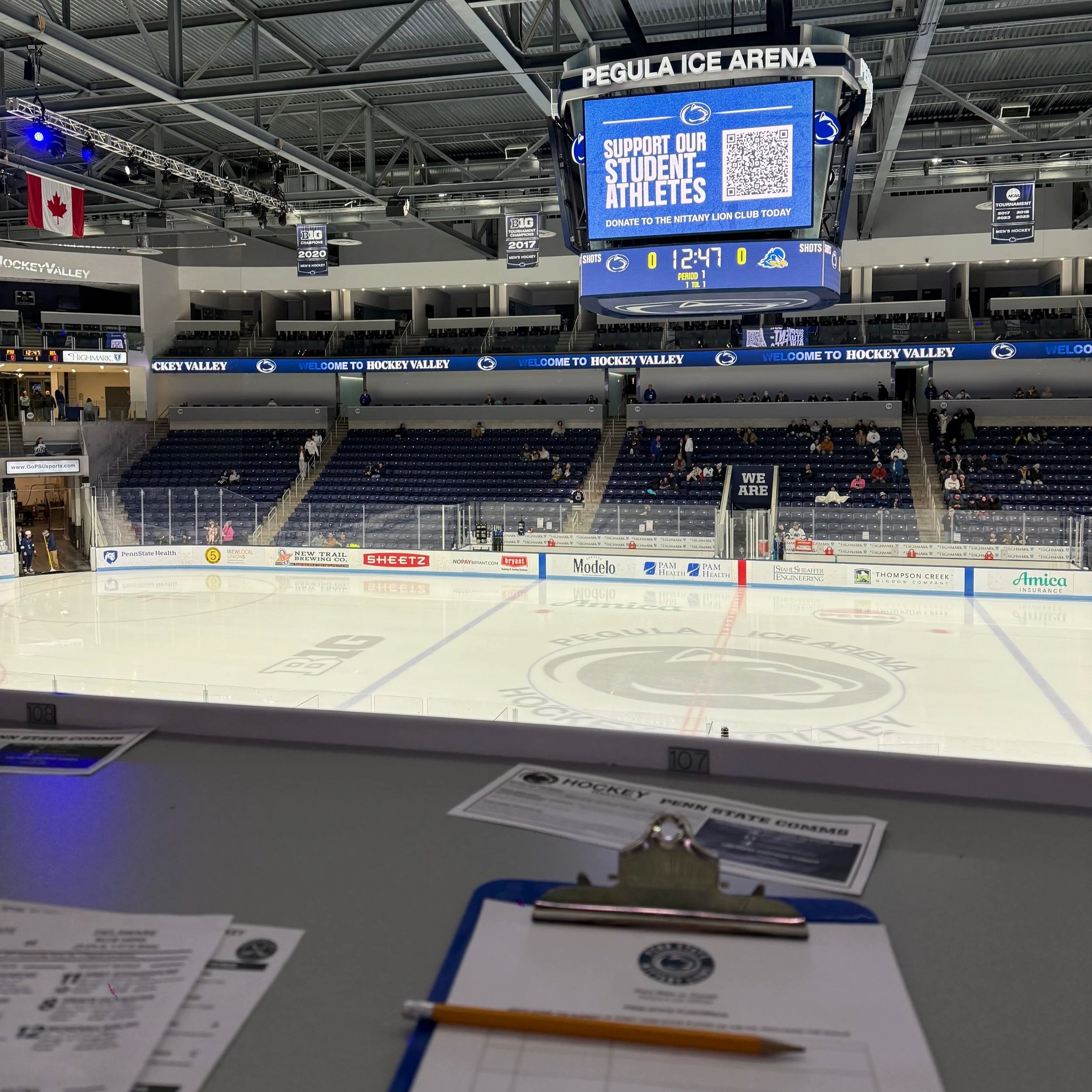 View of an ice hockey rink at Pegula Ice Arena with an empty seating area and a scoreboard displaying a support message for student athletes. The ice features the Penn State logo and the rink is surrounded by advertisements. The photo is taken from the scorer's table.
