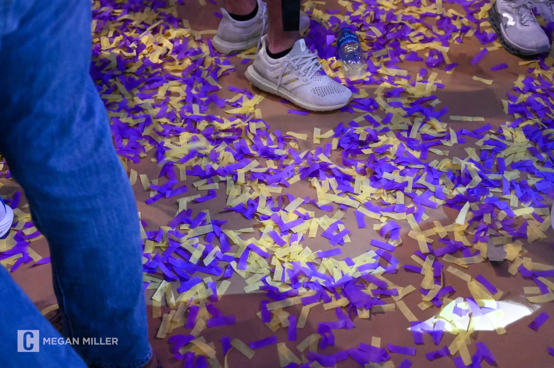 People standing on a floor covered with purple and yellow confetti, with some wearing athletic shoes and a plastic water bottle on the ground.
