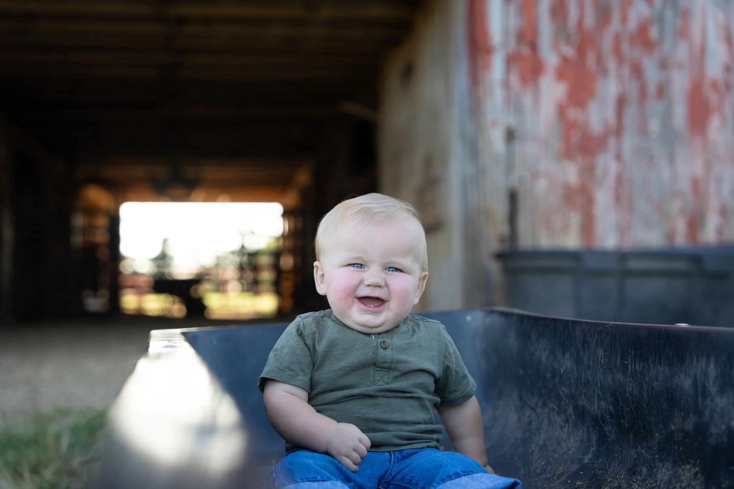 A young child with blonde hair and blue eyes sitting in a wheelbarrow, smiling and squinting outdoors with a rustic barn in the background.