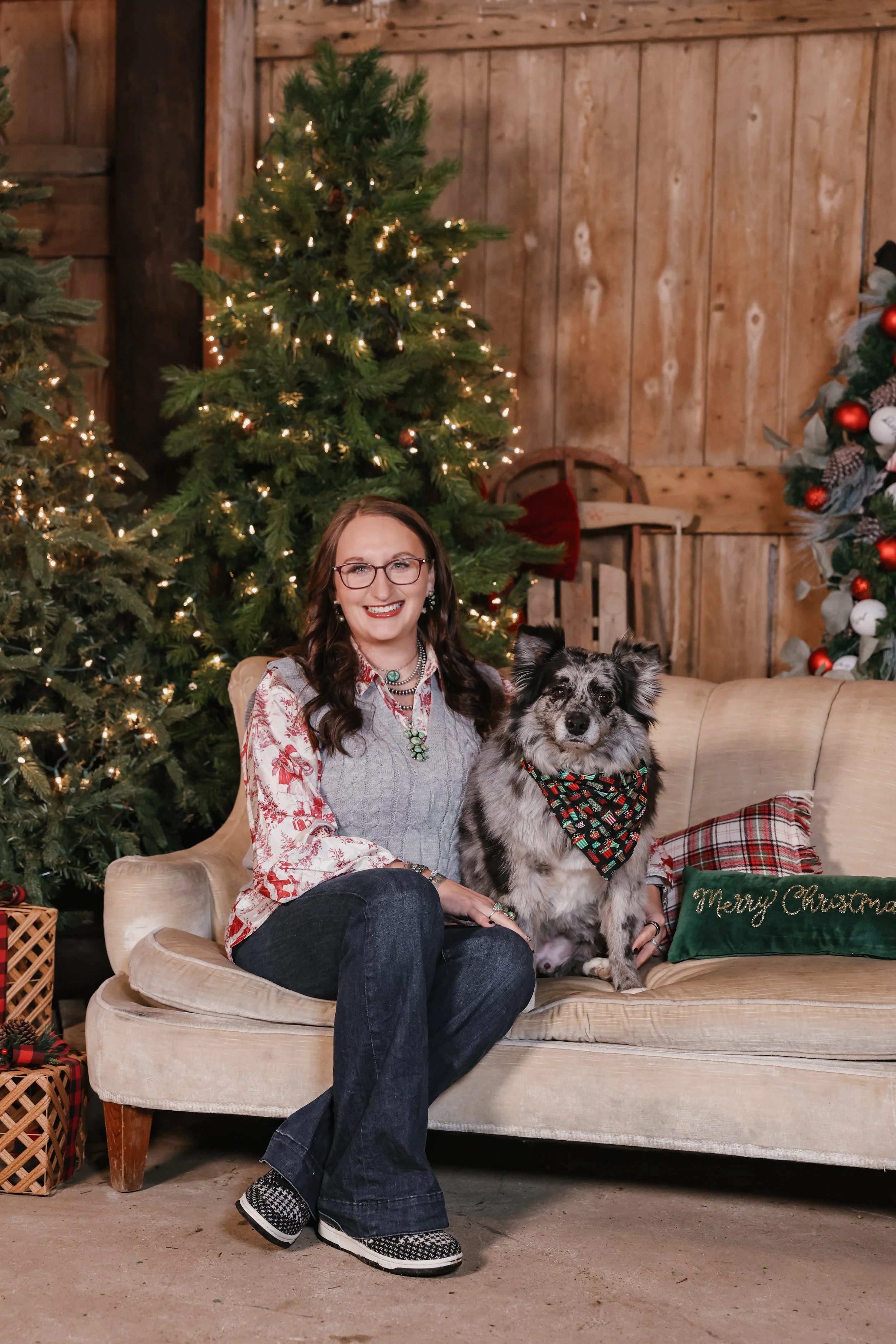 A woman sitting on a beige sofa next to a dog, in a Christmas setting with decorated Christmas trees and ornaments.
