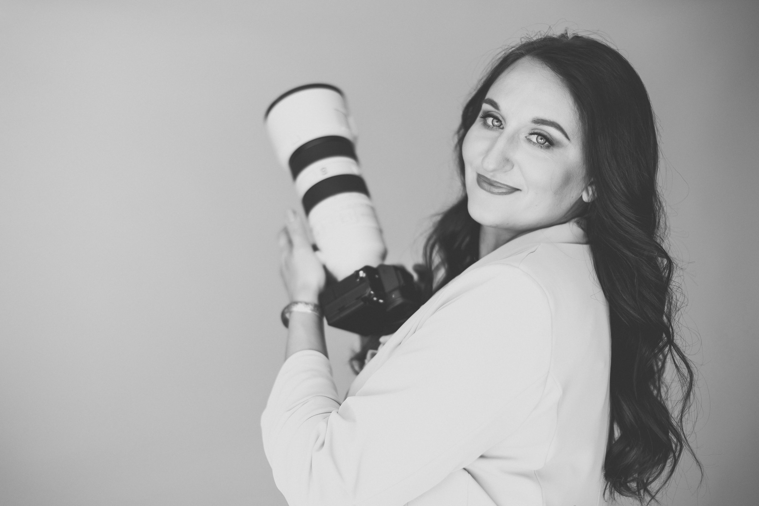 A woman with long wavy hair holding a camera with a large telephoto lens, looking at the camera with a slight smile in a black and white photo.