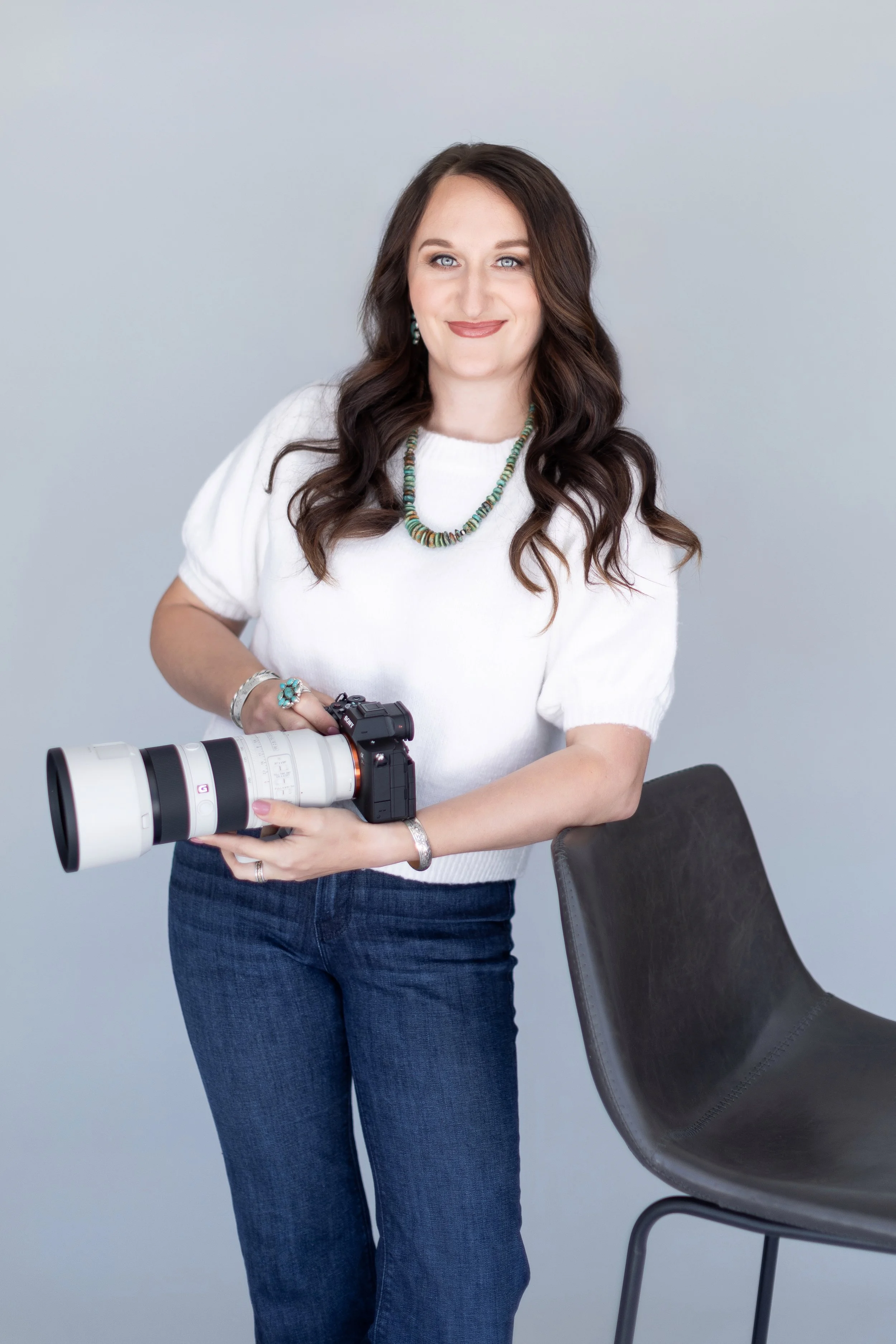 A woman holding a professional camera with a large telephoto lens, standing next to a black chair, against a plain light gray background.