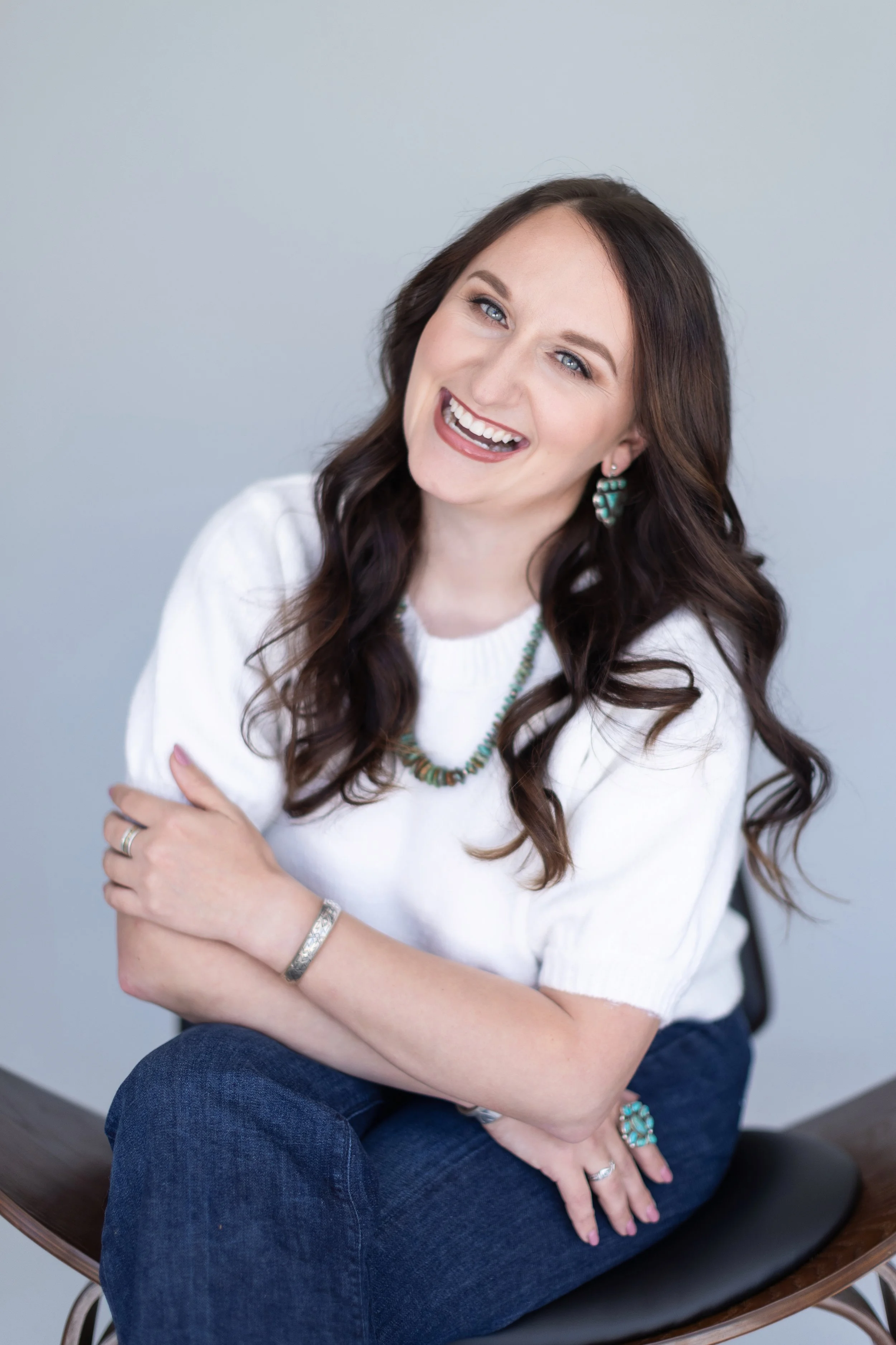 A woman with long brown hair and a bright smile sitting cross-legged on a wooden chair, wearing a white sweater, blue jeans, and turquoise jewelry, against a plain light gray backdrop.