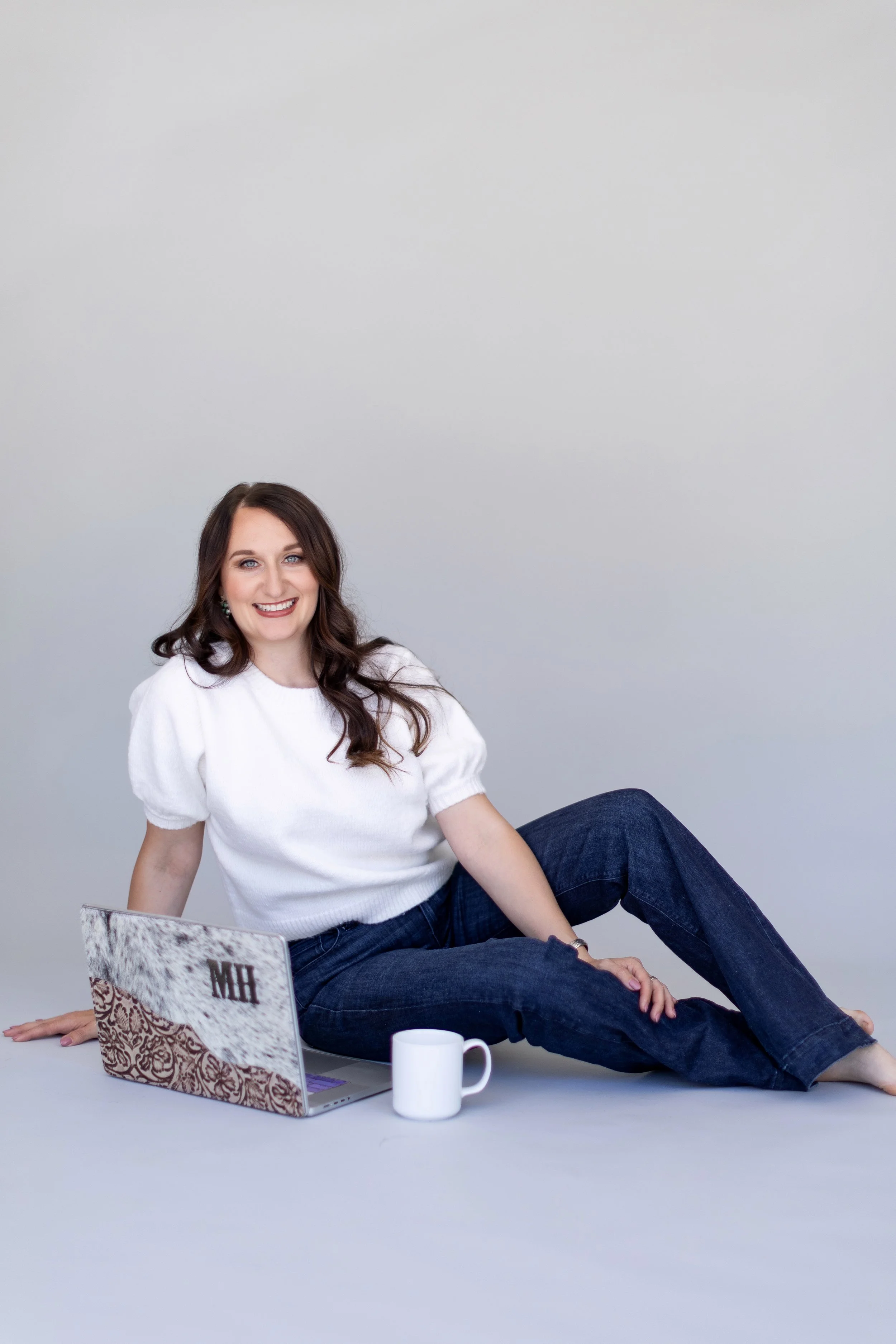 A woman with long brown hair, wearing a white sweater and dark jeans, sitting on the floor next to a laptop and a white mug, smiling at the camera against a plain light gray background.