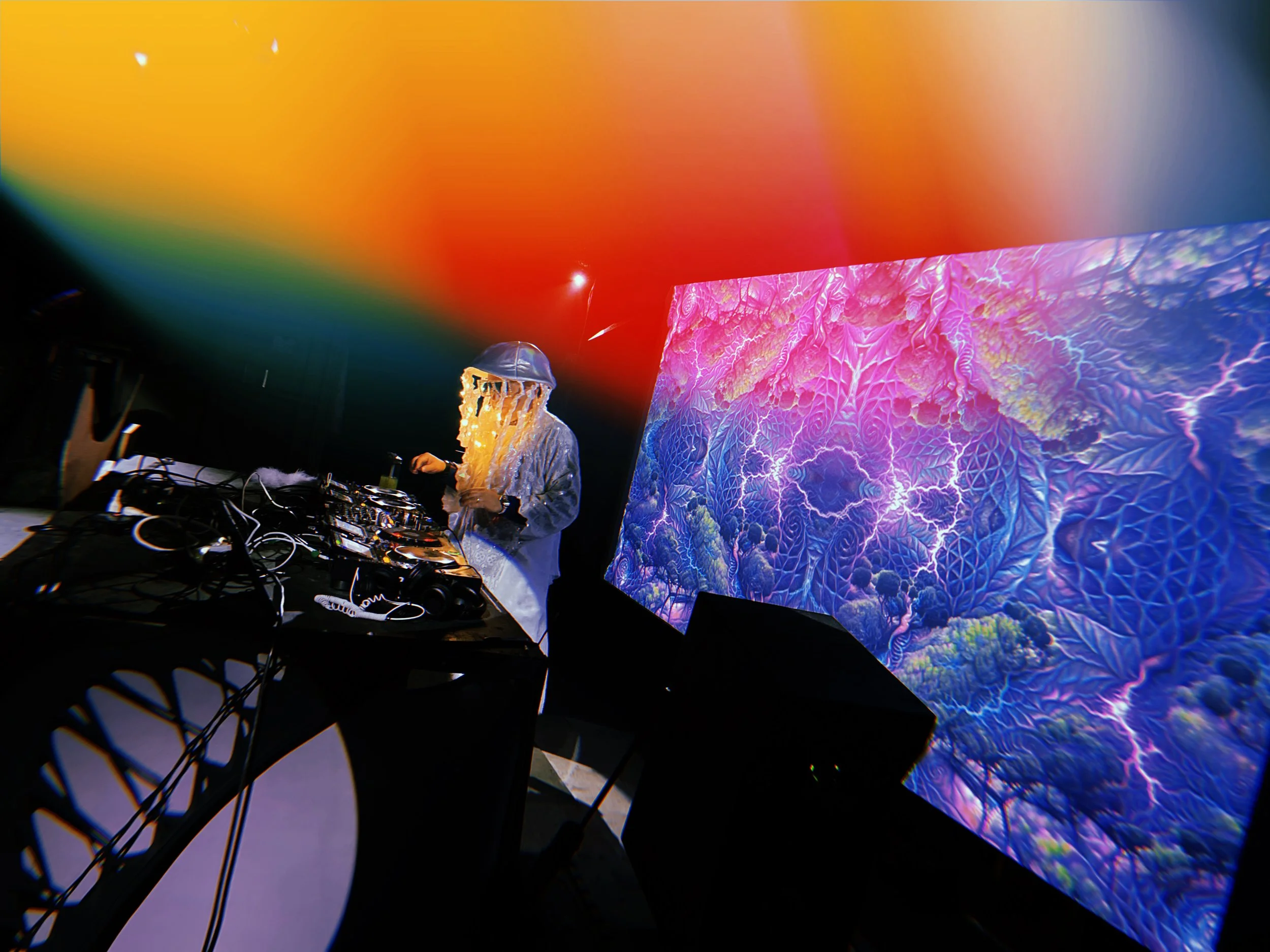 A DJ or performer with long blonde hair wearing headphones, standing in front of a large colorful digital display featuring vibrant, abstract graphics of lightning and intricate patterns, in a dark setting with a rainbow-colored light or projection above.