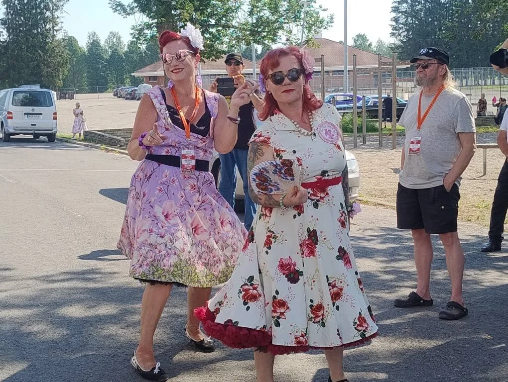 Kaksi naista pukeutunut retrotyylisesti, toinen hymyilee ja tekee rauhanmerkkiä, toinen katsoo vakavammin. Taustalla on muita ihmisiä ja autoja.