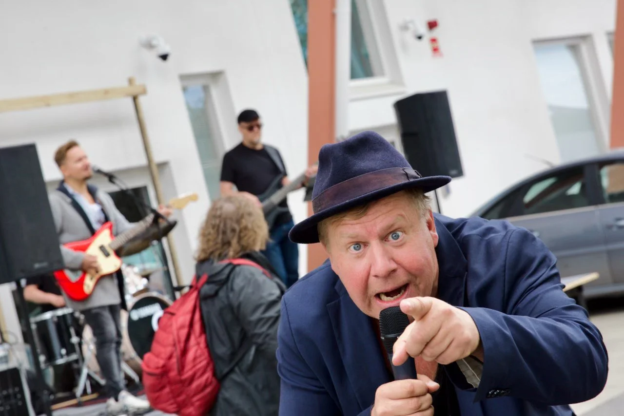 Mies esiintyy lavalla mikrofoni kädessä, ja taustalla on bändi musiikkiesitystä varten. Kuva Liperi Truck Meetingista 2025.