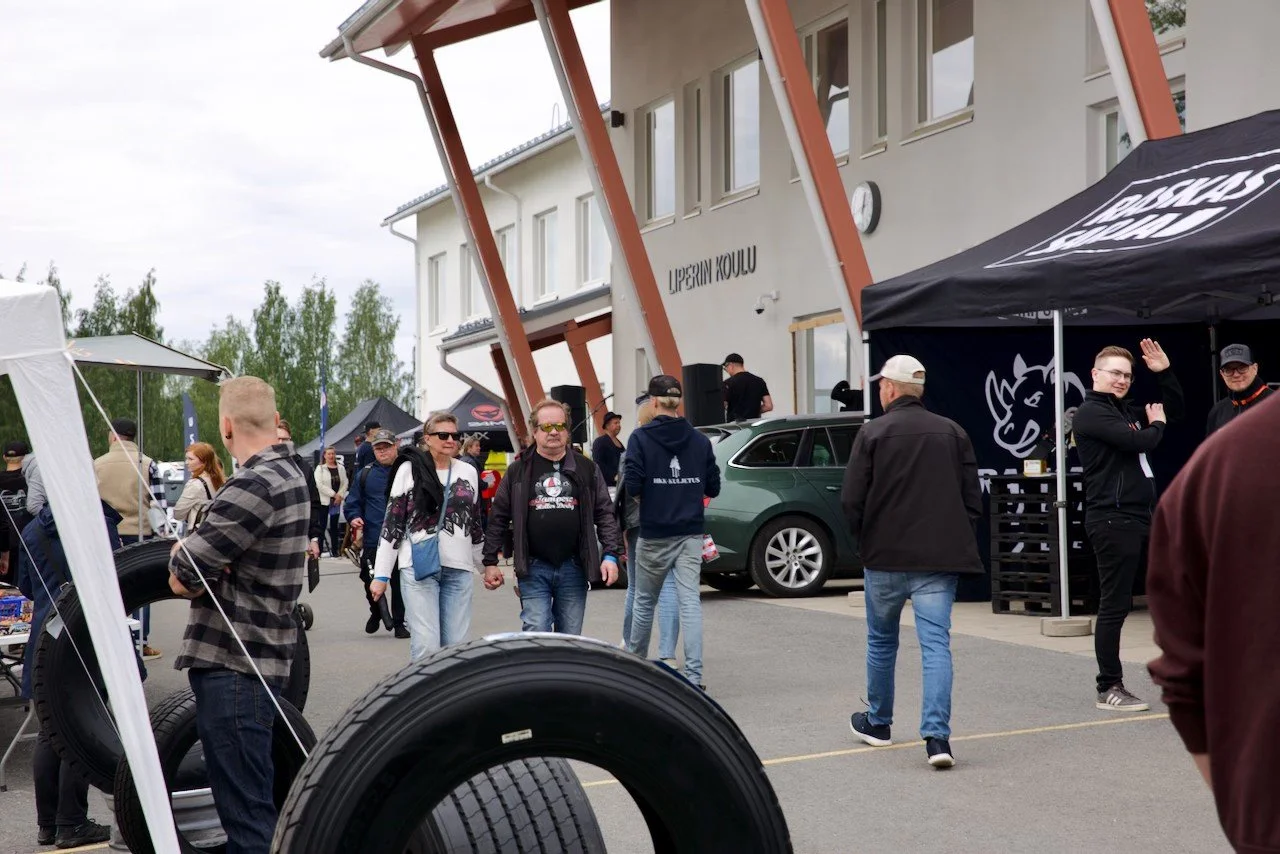 Ihmisiä tapahtumassa ulkona, myyntikojuja ja autoja, rakennus taustalla, sään sää. Tapahtuma liittyy autoharrastukseen.