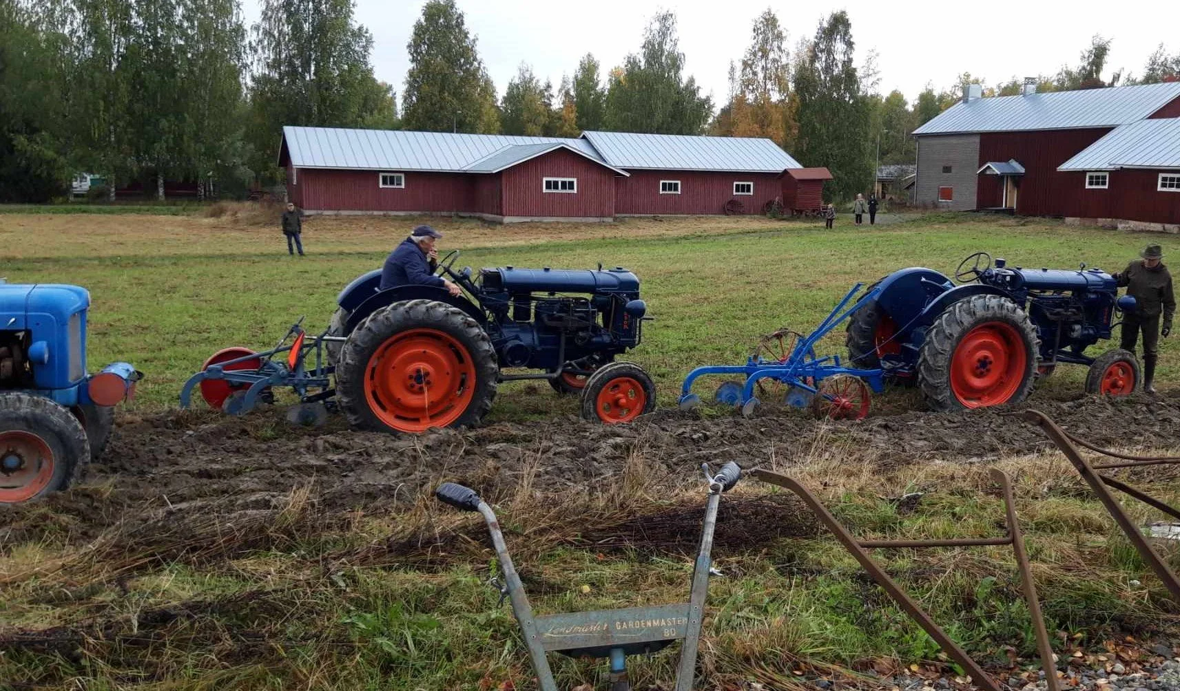 Kaksi vanhanaikaista traktoria, yksi mies istuu ja toinen seisoo, työskentelee peltoa alasajossa. Taustalla on punaisia ja harmaita maatilarakennuksia ja ihmisiä katsomassa. Kuva Hakoja, Tonkkia ja Veteraanikoneita -tapahtumasta 2025.