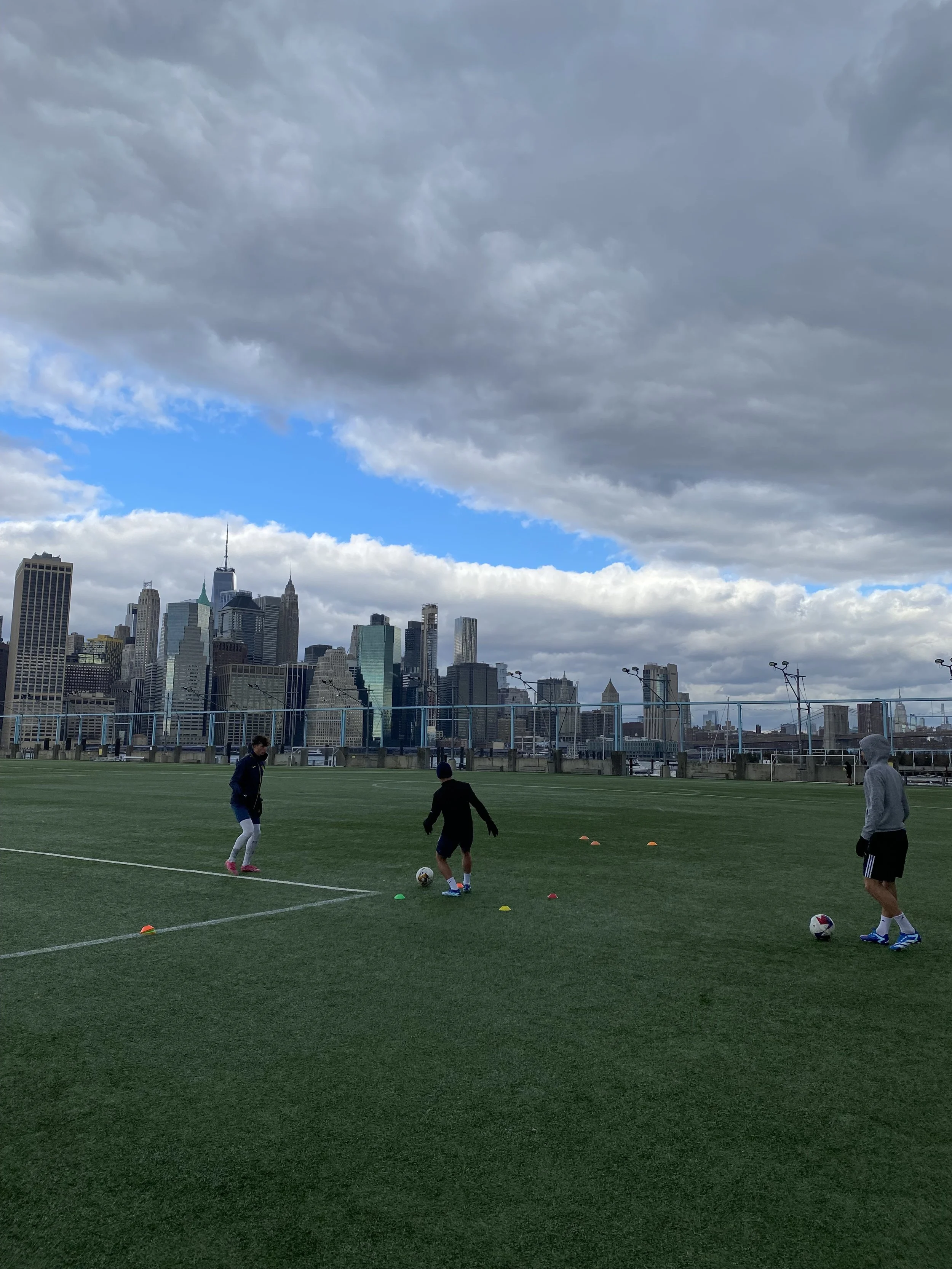 Three people on a soccer field practicing with cones and soccer balls under cloudy sky, with a city skyline in the background.