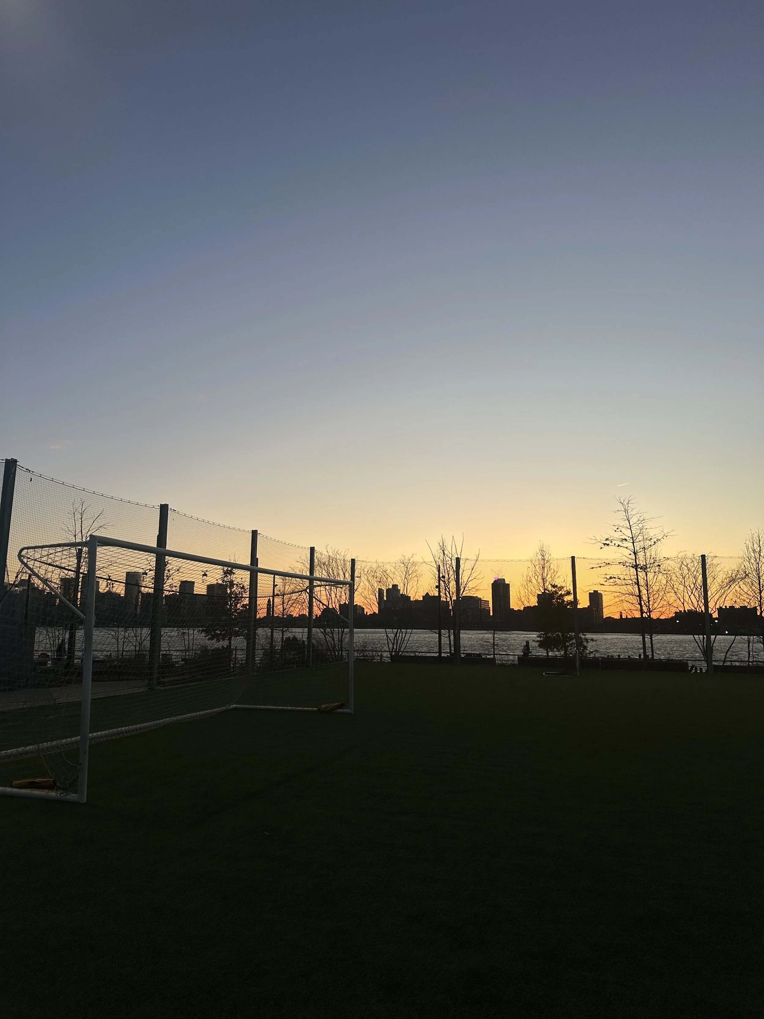Soccer goal on a grassy field near a river during sunset, with city skyline in the distance and trees along the riverbank.