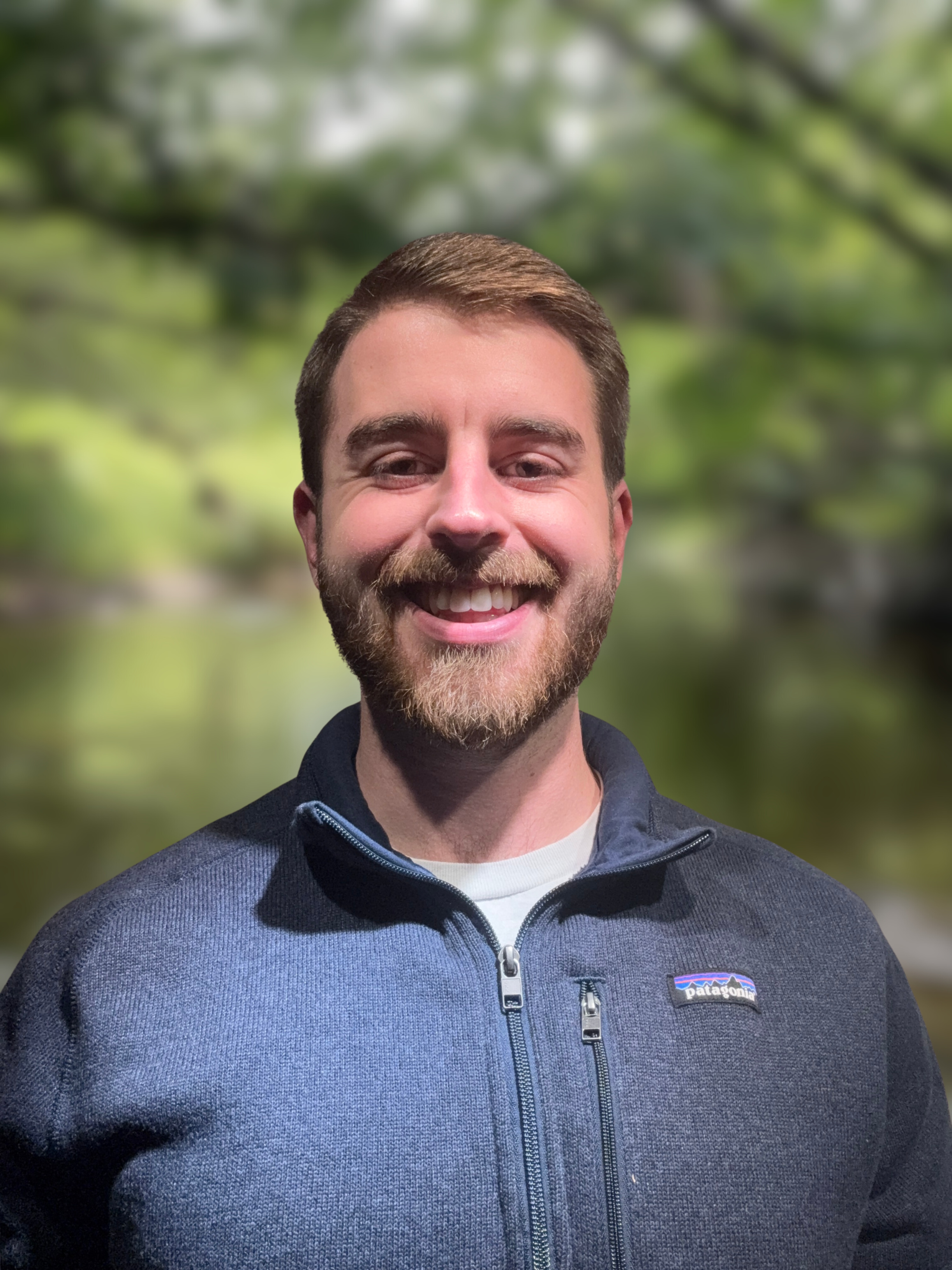 A smiling man with short brown hair, a beard, dressed in a dark blue Patagonia jacket, standing outdoors with blurry green trees in the background.