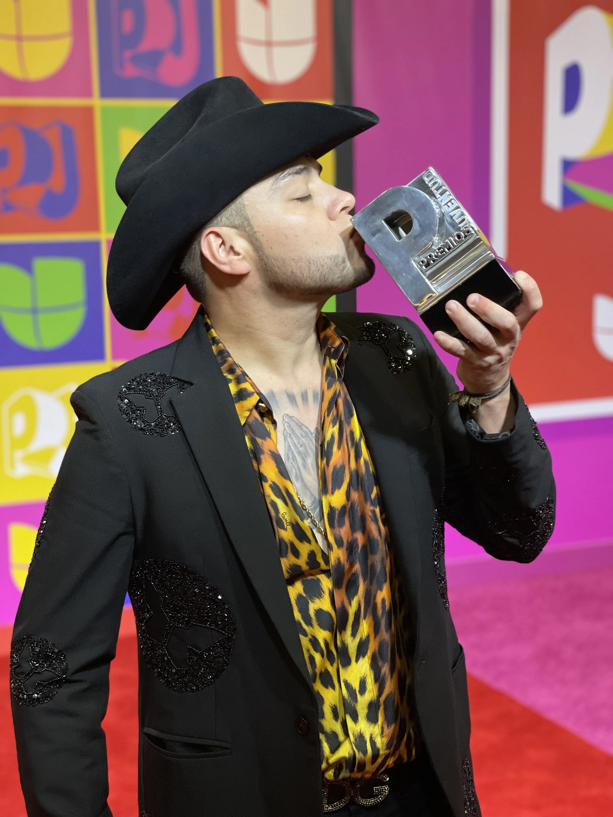 A man wearing a black cowboy hat and leopard print shirt, holding a large metallic P de Oscar statue and kissing it, standing in front of a colorful background with multiple Oscar logos.
