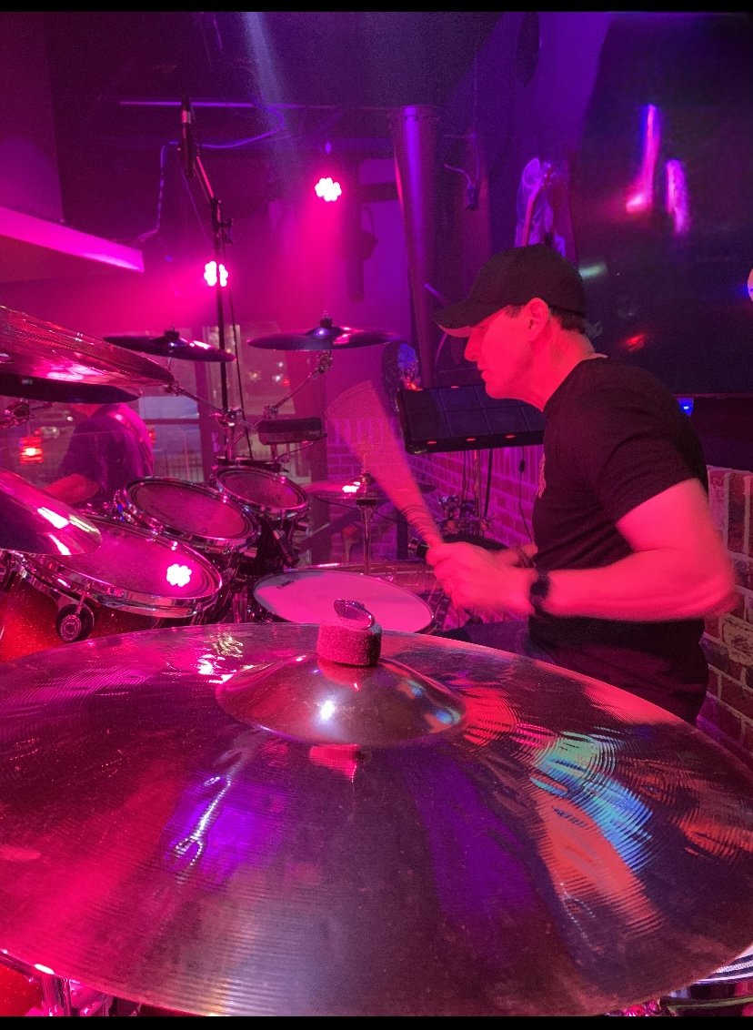 A person playing acoustic drums in a dimly lit venue with purple and pink stage lights.