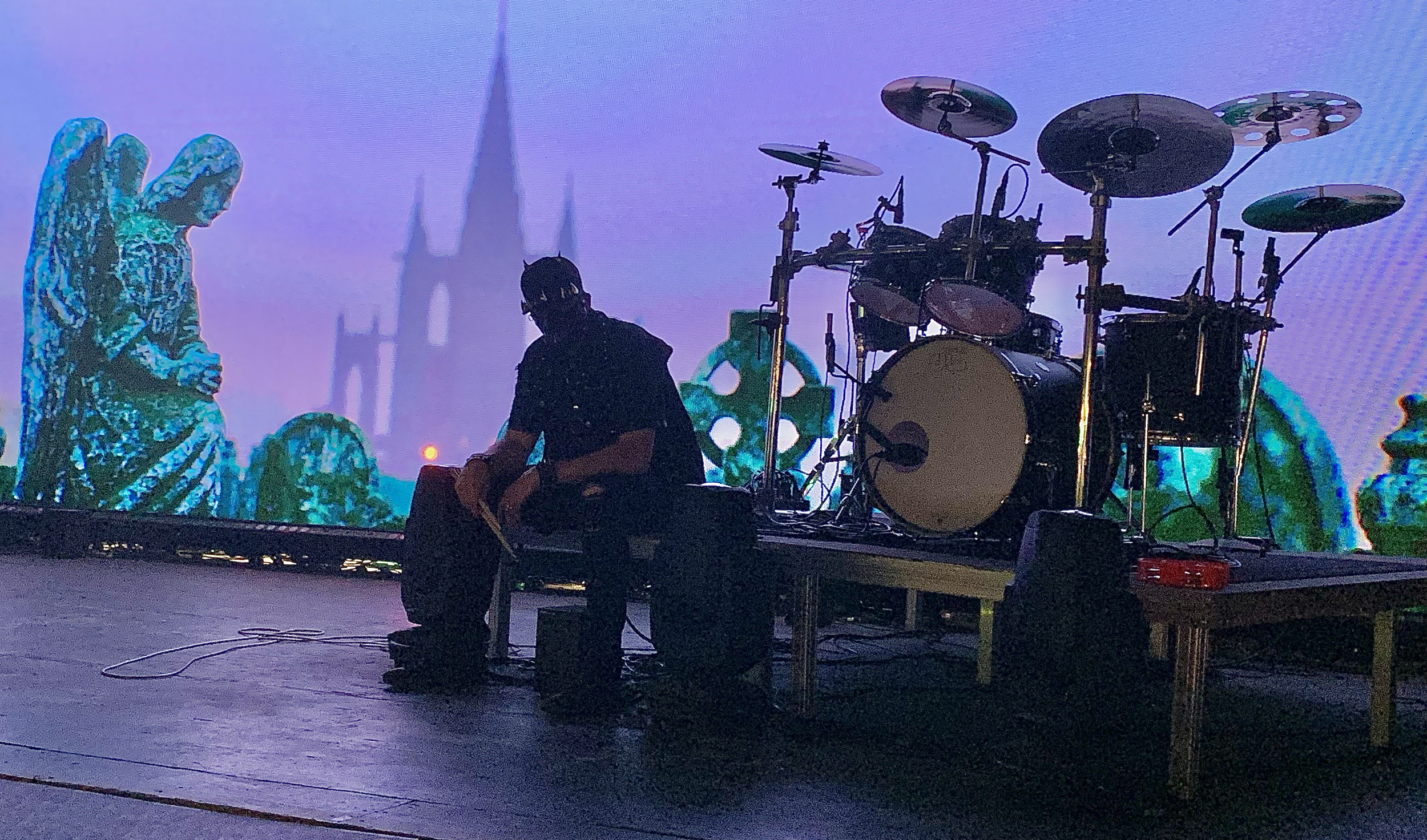 Silhouette of a person sitting next to a drum set on a stage with animated background visuals depicting a cathedral and turrets.