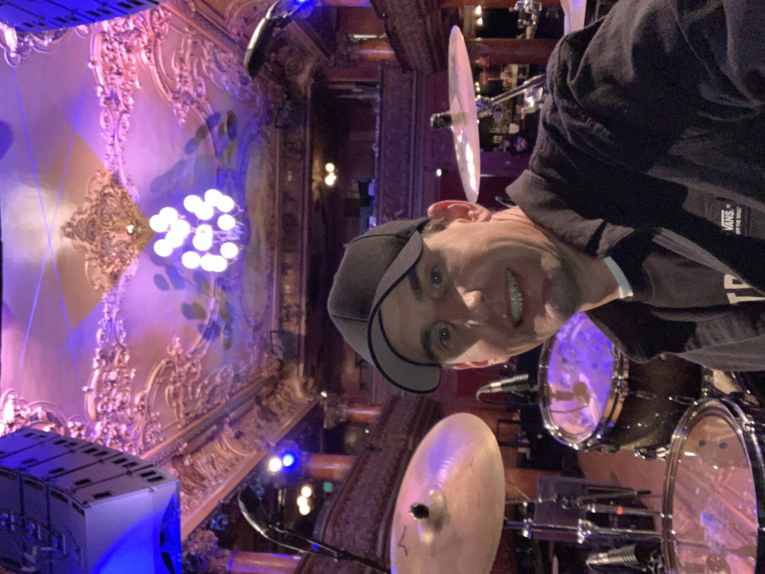 A man wearing a black shirt and a black cap taking a selfie in a room with ornate, gold-colored ceiling decorations and drums. The room is dimly lit with purple and pink lighting.