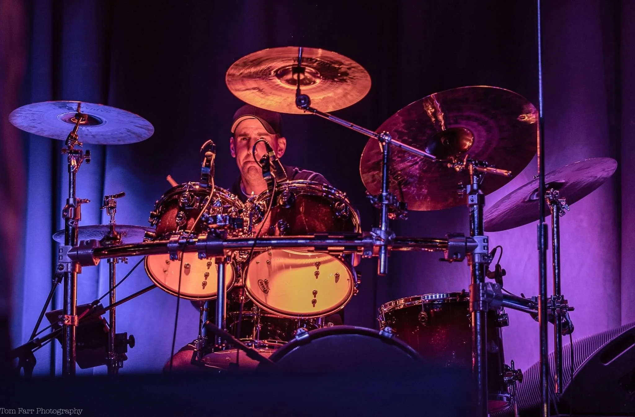 A man playing a drum set on stage with purple and blue lighting.