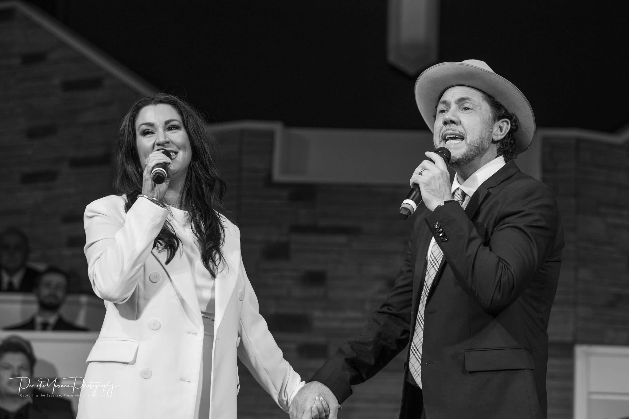 Two singers, a woman in a light-colored blazer and a man in a dark suit and hat, hold hands and sing into microphones on stage, with an audience in the background.