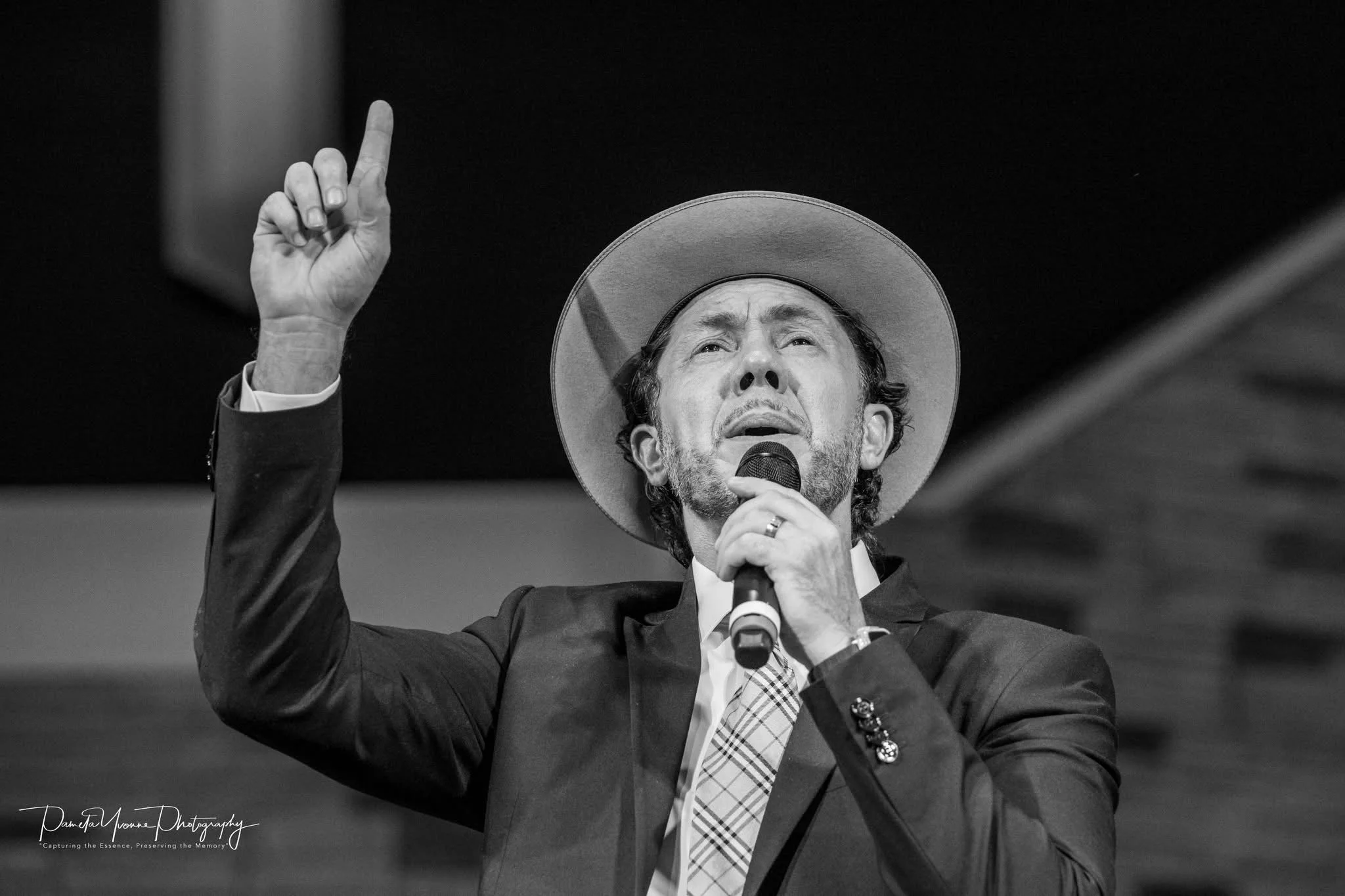 A man in a suit and hat passionately speaking or singing into a microphone, raising his right hand with the index finger pointing upward.